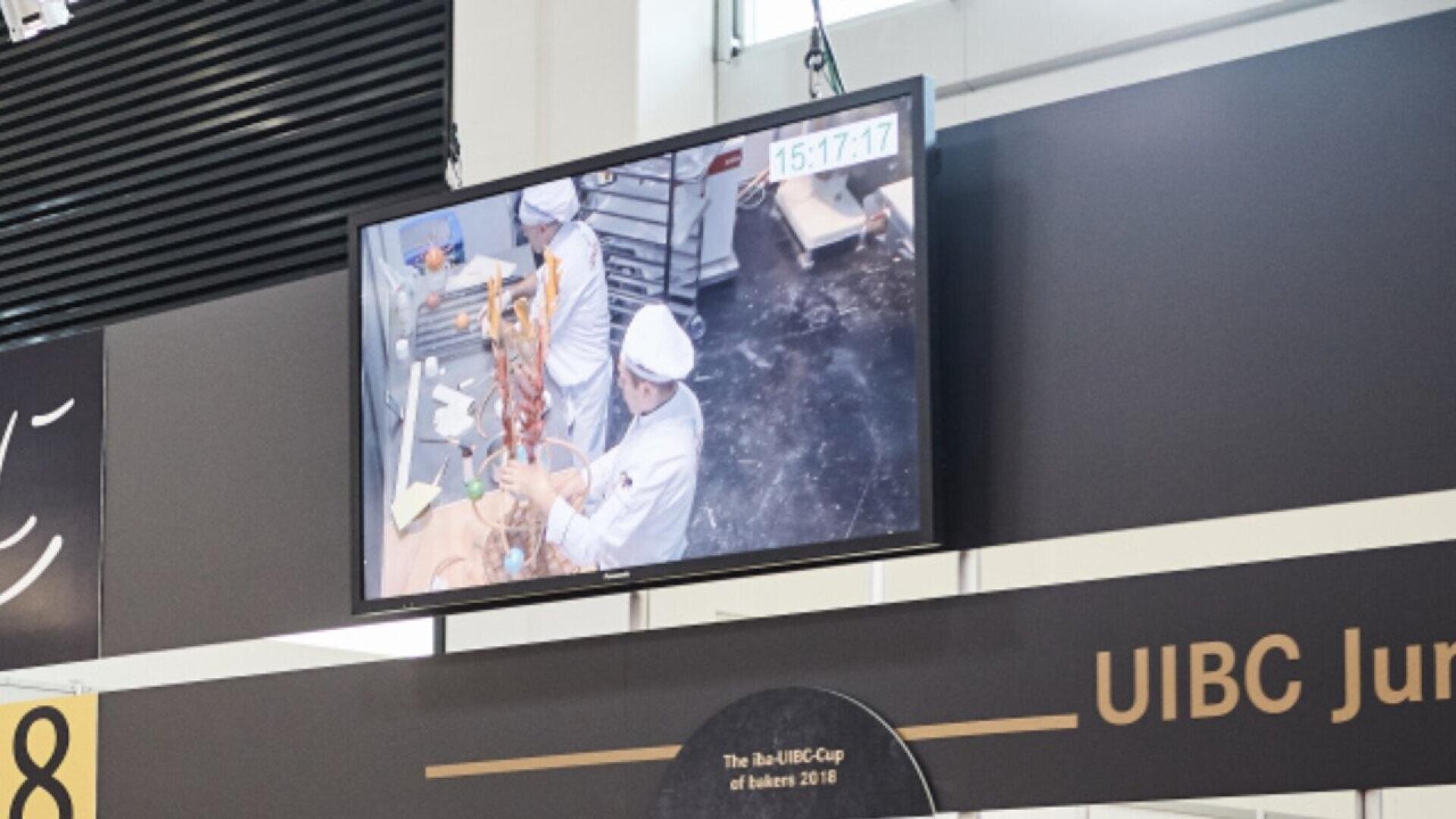 A monitor displays a live feed of bakers in white uniforms assembling a tall pastry structure in a kitchen, with a digital clock showing the time 15:17:17. The scene is part of the UIBC Junior Cup event.