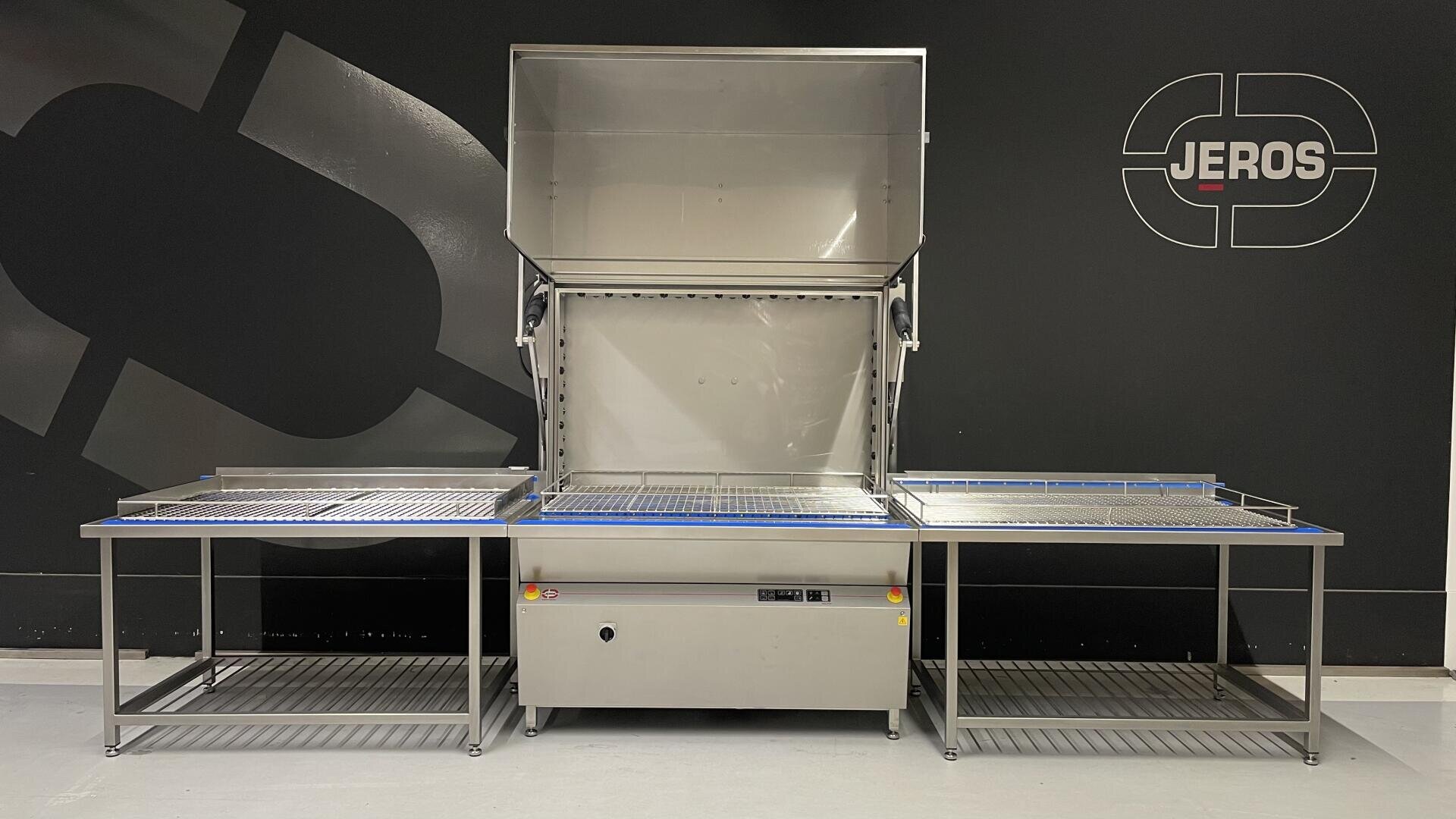 A large stainless steel industrial cleaning machine with an open lid, side tables on both sides, and the brand name JEROS displayed on a black wall in the background.