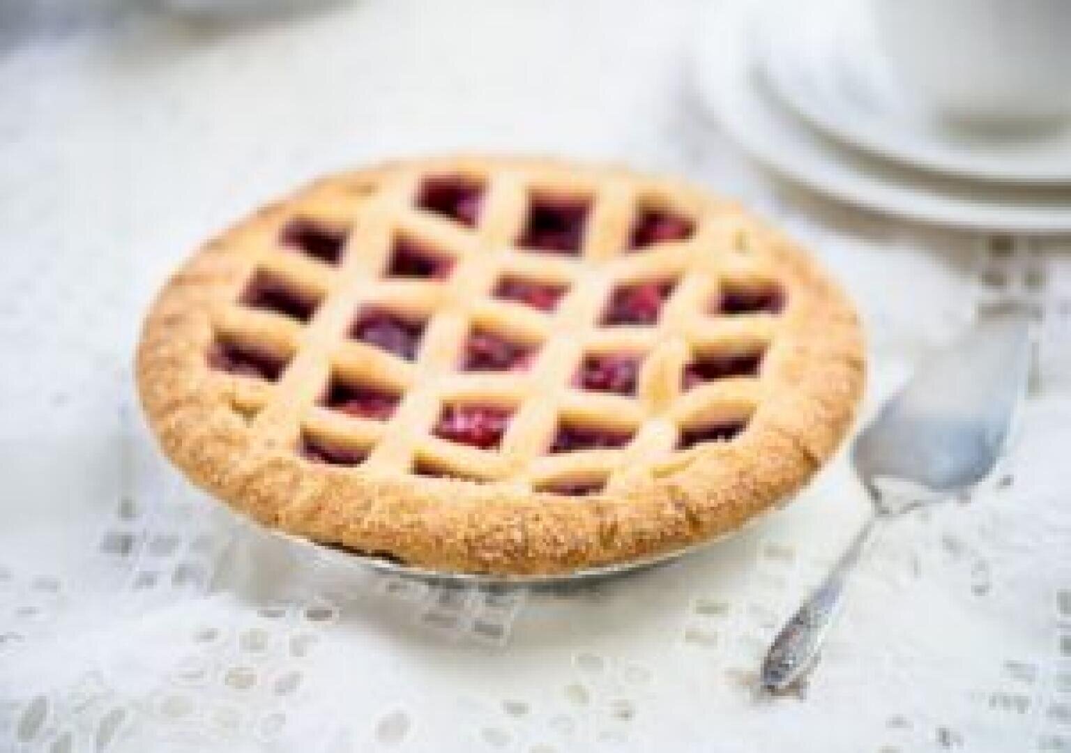 A freshly baked pie with a golden lattice crust filled with red fruit sits on a lace tablecloth beside a silver pie server and a white cup and saucer.