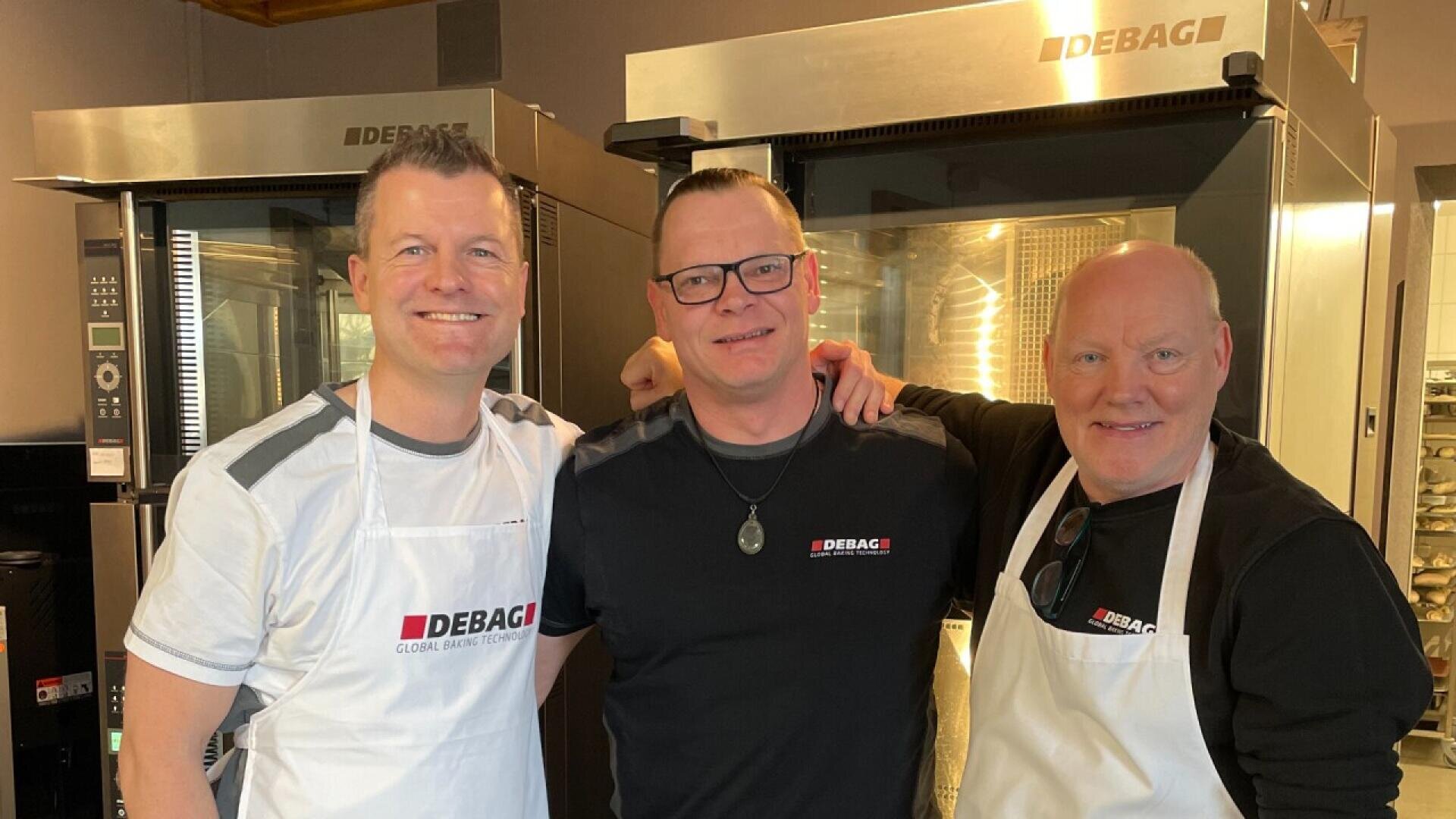 Three men wearing DEBAG-branded shirts and aprons stand smiling together in a bakery kitchen, with industrial ovens visible in the background.
