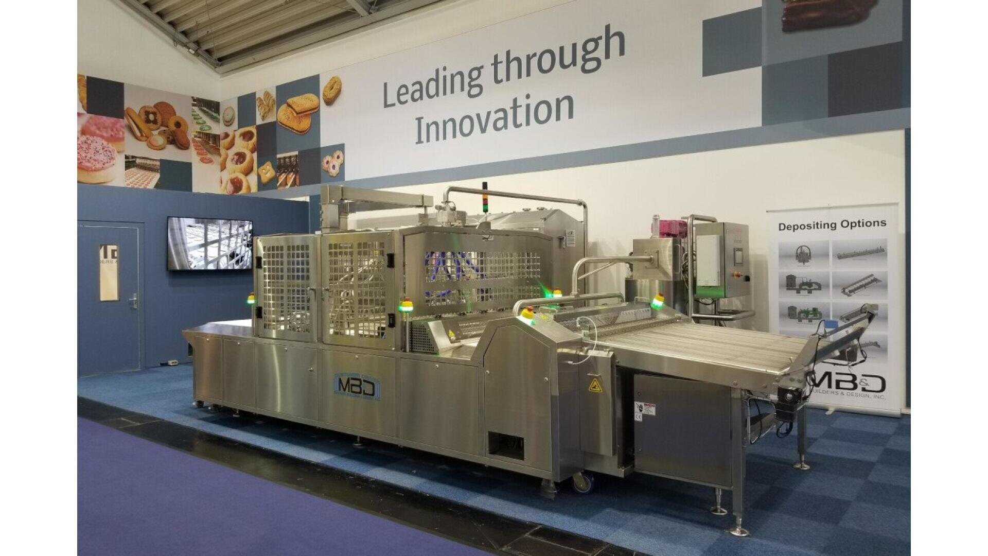 Eine große industrielle Lebensmittelverarbeitungsmaschine steht in einem Messestand mit Schildern, auf denen "Leading through Innovation" steht, und Bildern von Backwaren an der Wand im Hintergrund.