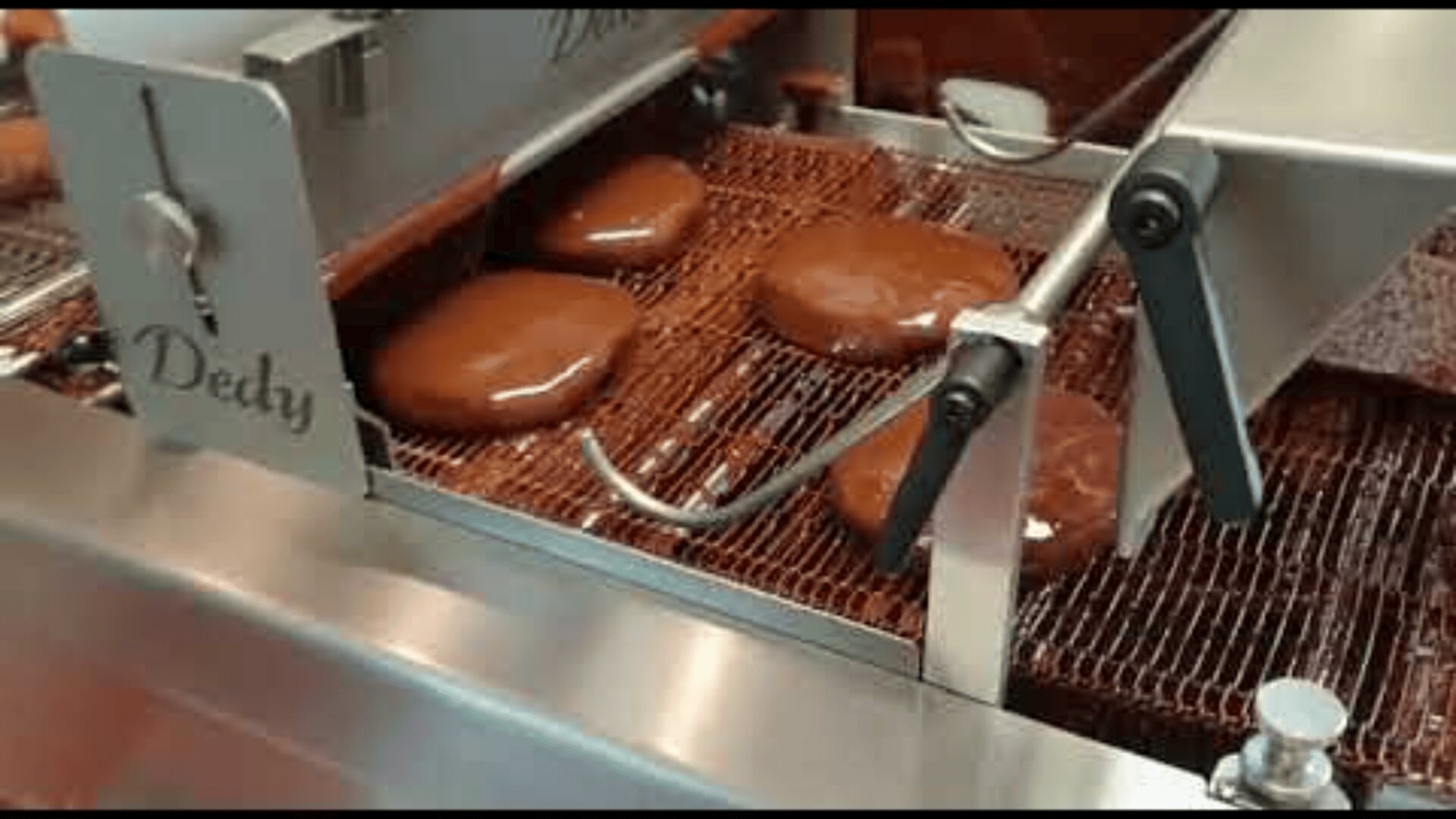 Chocolate-coated pastries move along a conveyor belt inside a machine, being enrobed in liquid chocolate as part of an automated production process.
