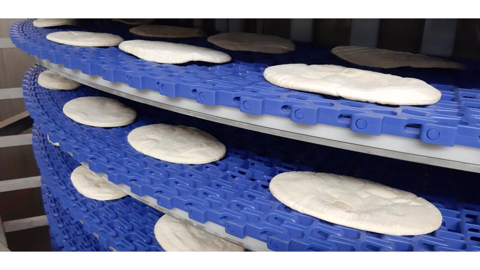 Flatbreads are placed on multiple levels of a blue conveyor belt inside an industrial bakery, likely as part of an automated production or cooling process.