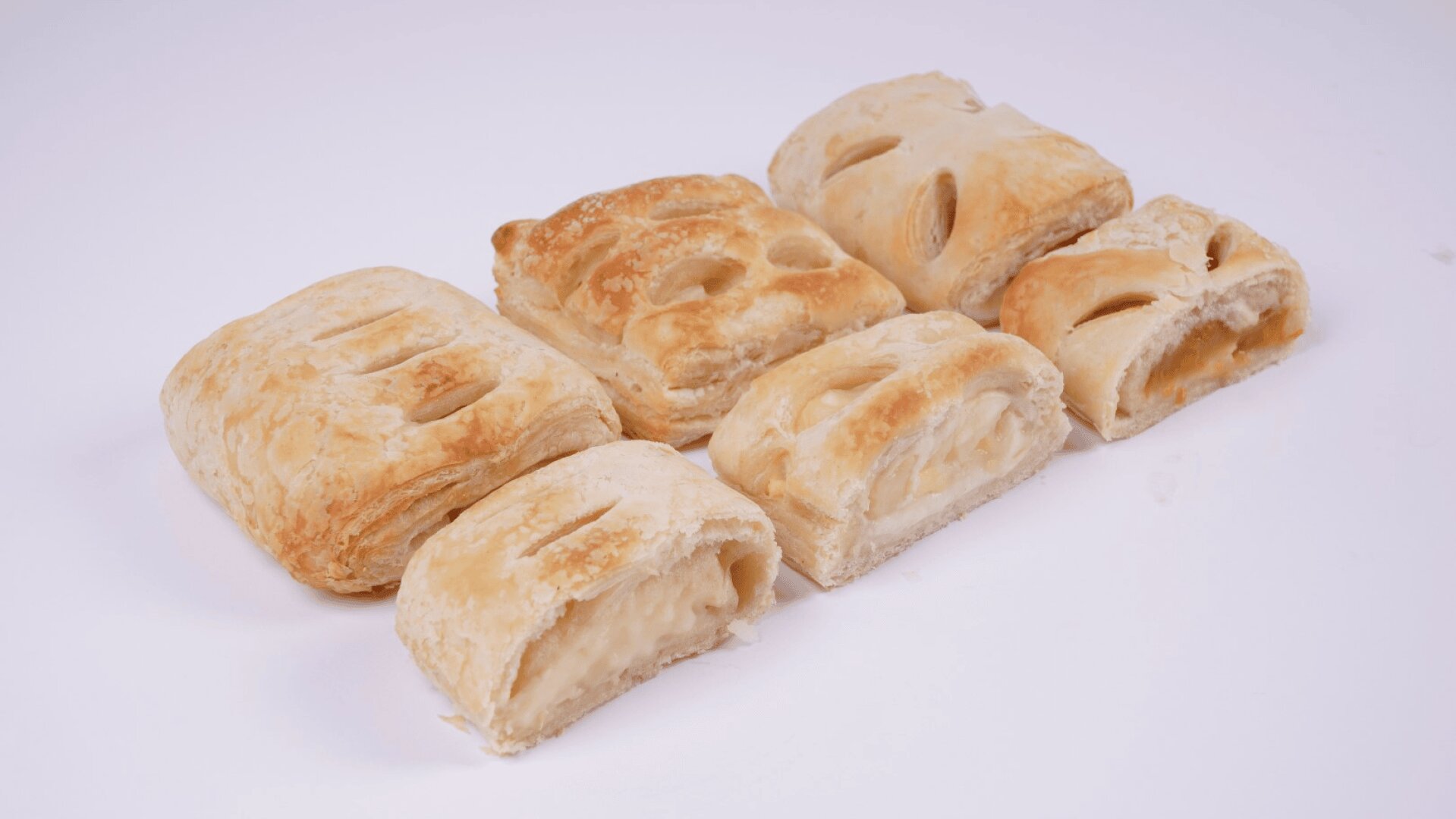 Sechs flockige, goldbraune Gebäcke mit sichtbaren Füllungen sind auf einer weißen Fläche angeordnet. Einige Gebäckstücke sind aufgeschnitten und geben den Blick auf cremige und fruchtige Füllungen frei.