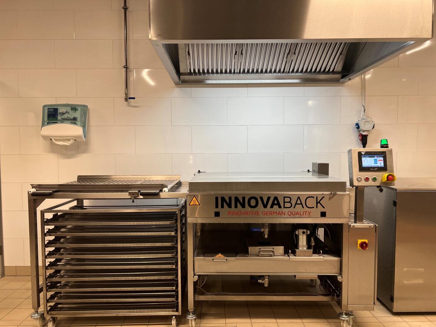 Eine Großküche ist mit Geräten aus Edelstahl ausgestattet, darunter ein großer Backofen mit der Aufschrift INNOVABACK und einem digitalen Bedienfeld, ein Metallgestell mit Tabletts, eine Abzugshaube und ein an der Wand montierter Papierhandtuchspender.