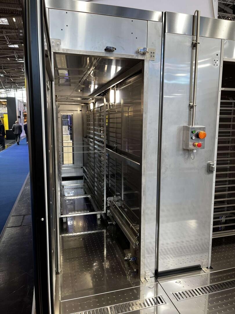 Ein großer, industrieller Edelstahlofen oder Gärraum mit offenen Türen, Einschüben im Inneren und einem Bedienfeld mit roten und grünen Tasten auf der rechten Seite. Die Umgebung erinnert an eine Messe oder Ausstellungshalle.