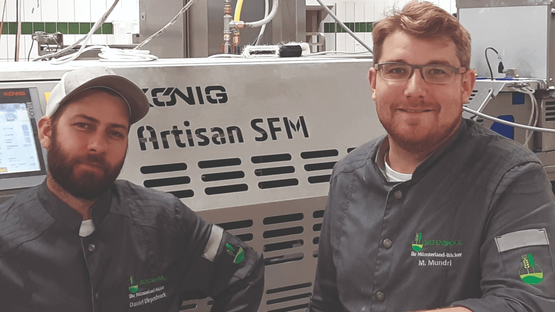 Zwei Männer in grauen Kochjacken und Mützen stehen vor einer großen Industriemaschine mit der Aufschrift KÖNIG Artisan SFM. Sie lächeln und scheinen sich in einer professionellen Küche oder Bäckerei zu befinden.