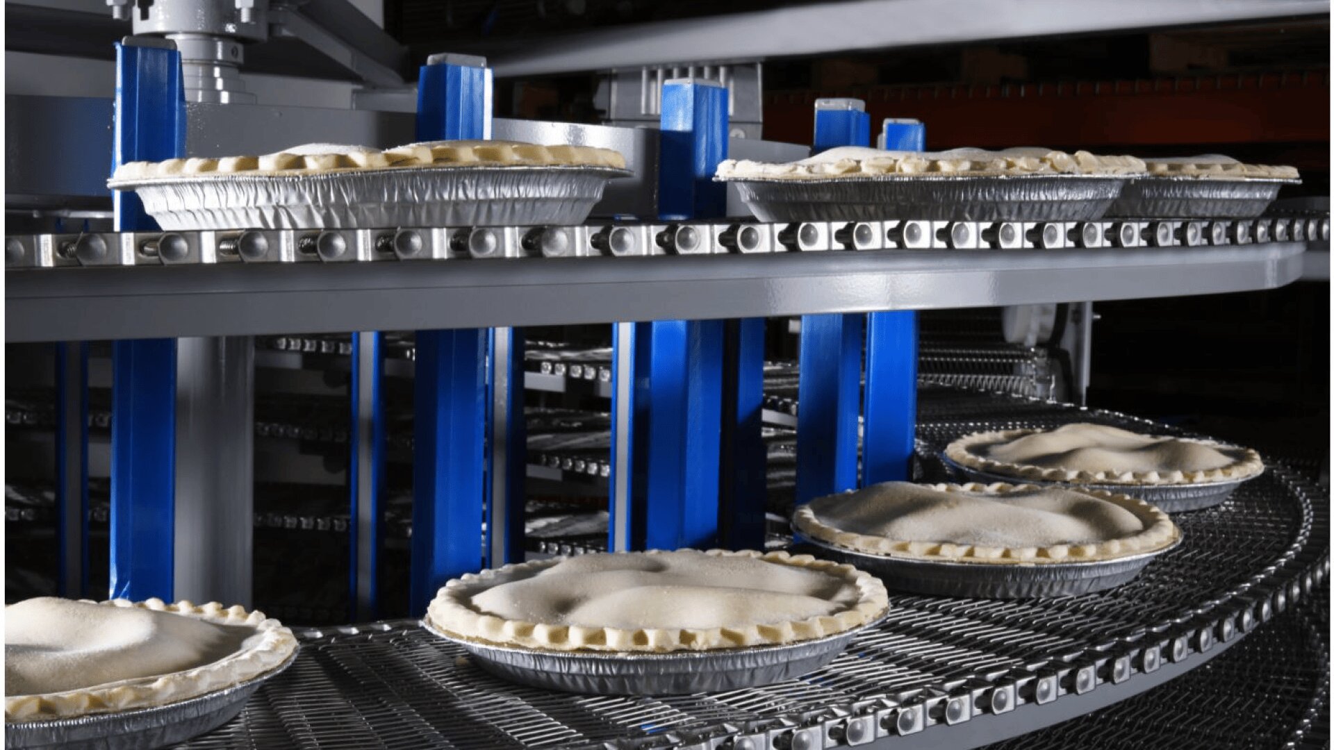 Multiple unbaked pies in metal pans move along a spiral conveyor belt in an industrial bakery setting, with blue vertical supports and a metallic wire mesh surface visible.