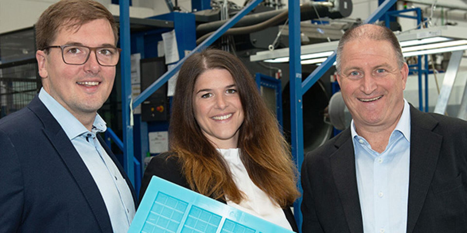 Drei lächelnde Menschen in Geschäftskleidung stehen in einem Haus und halten eine blaue Plastikform. Im Hintergrund sind Industriemaschinen und Metallkonstruktionen zu sehen.