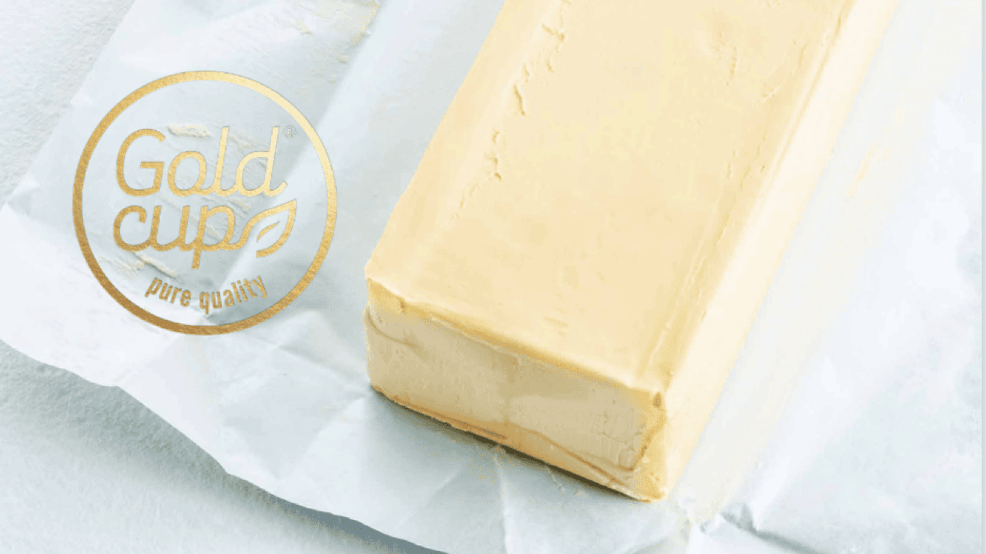 A rectangular block of butter sits on white parchment paper. A gold circular Gold Cup pure quality logo with a leaf design is visible on the left side of the image.