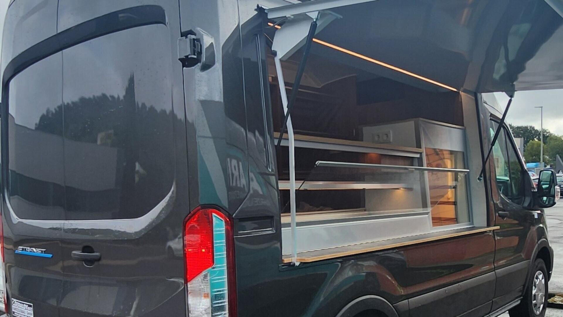 A black food truck with its side window open, revealing an empty serving counter and shelves inside, parked in an outdoor area on a cloudy day.