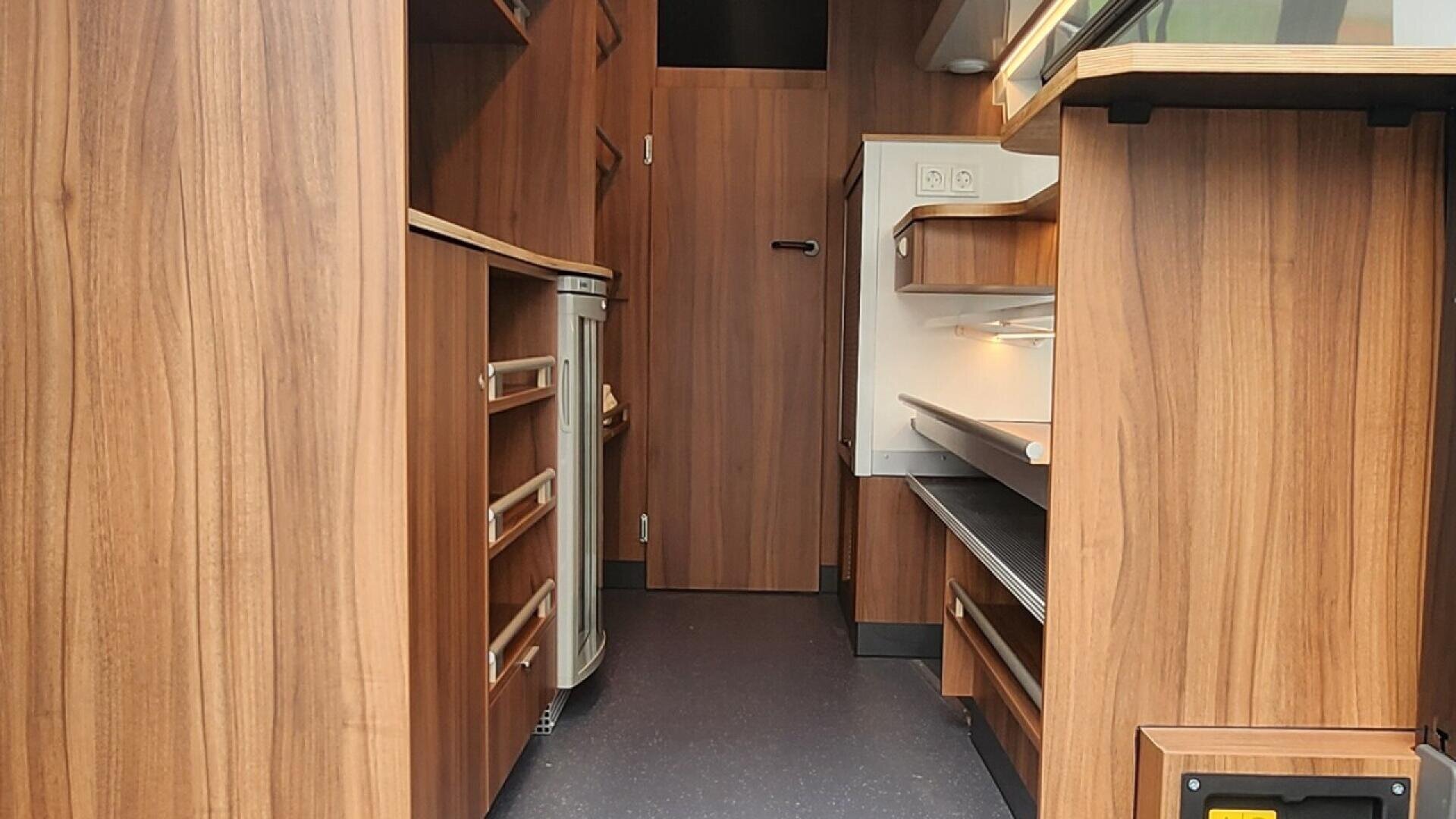 Interior of a modern camper van featuring wood paneling, open shelves, cabinets, a countertop, and a door at the back; soft lighting creates a cozy atmosphere.