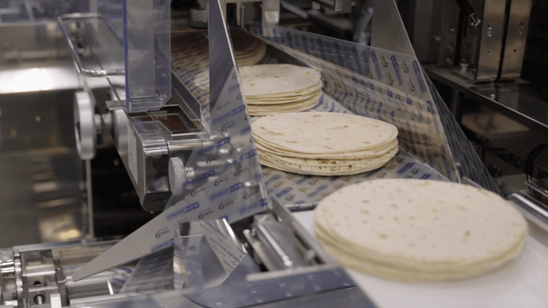 Stacks of flour tortillas move along a conveyor belt in a food processing factory, with packaging materials and stainless steel machinery visible.