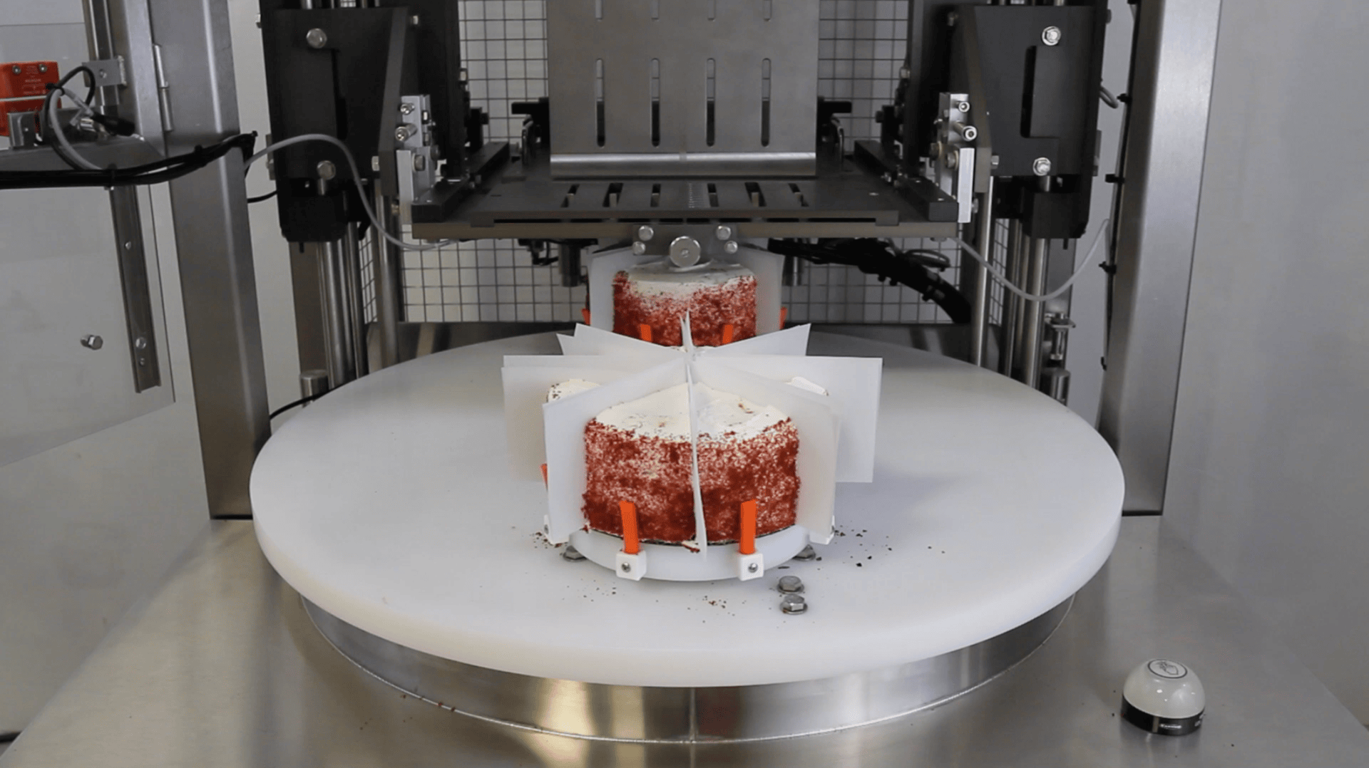 A red velvet cake secured in a white cake slicer machine, positioned on a round platform, ready to be evenly cut by metal blades within a stainless steel device.