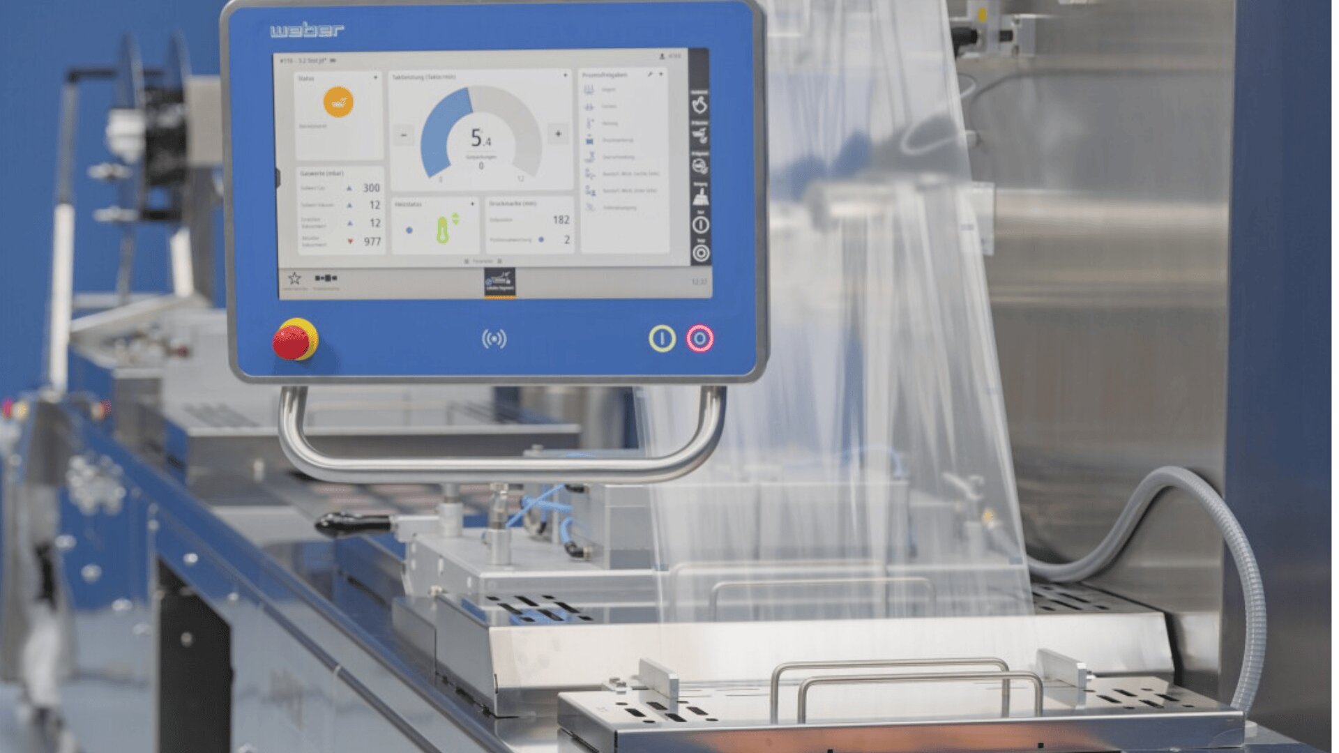A close-up of a blue industrial control panel with a touchscreen display showing graphs and data, mounted on packaging machinery with transparent plastic film in a factory setting.