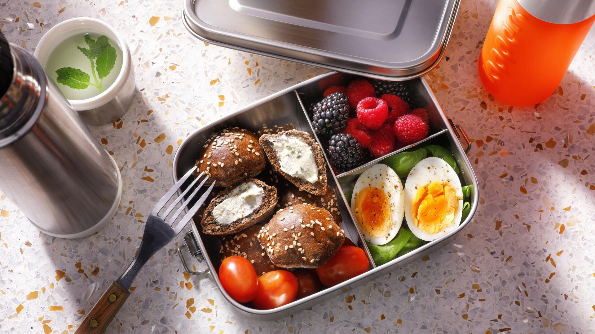 A lunchbox with sesame-topped stuffed mushrooms, cherry tomatoes, boiled egg halves with pepper, blackberries, raspberries, blueberries, and spinach. Nearby are a fork, a drink flask, an orange bottle, and a cup with mint leaves.