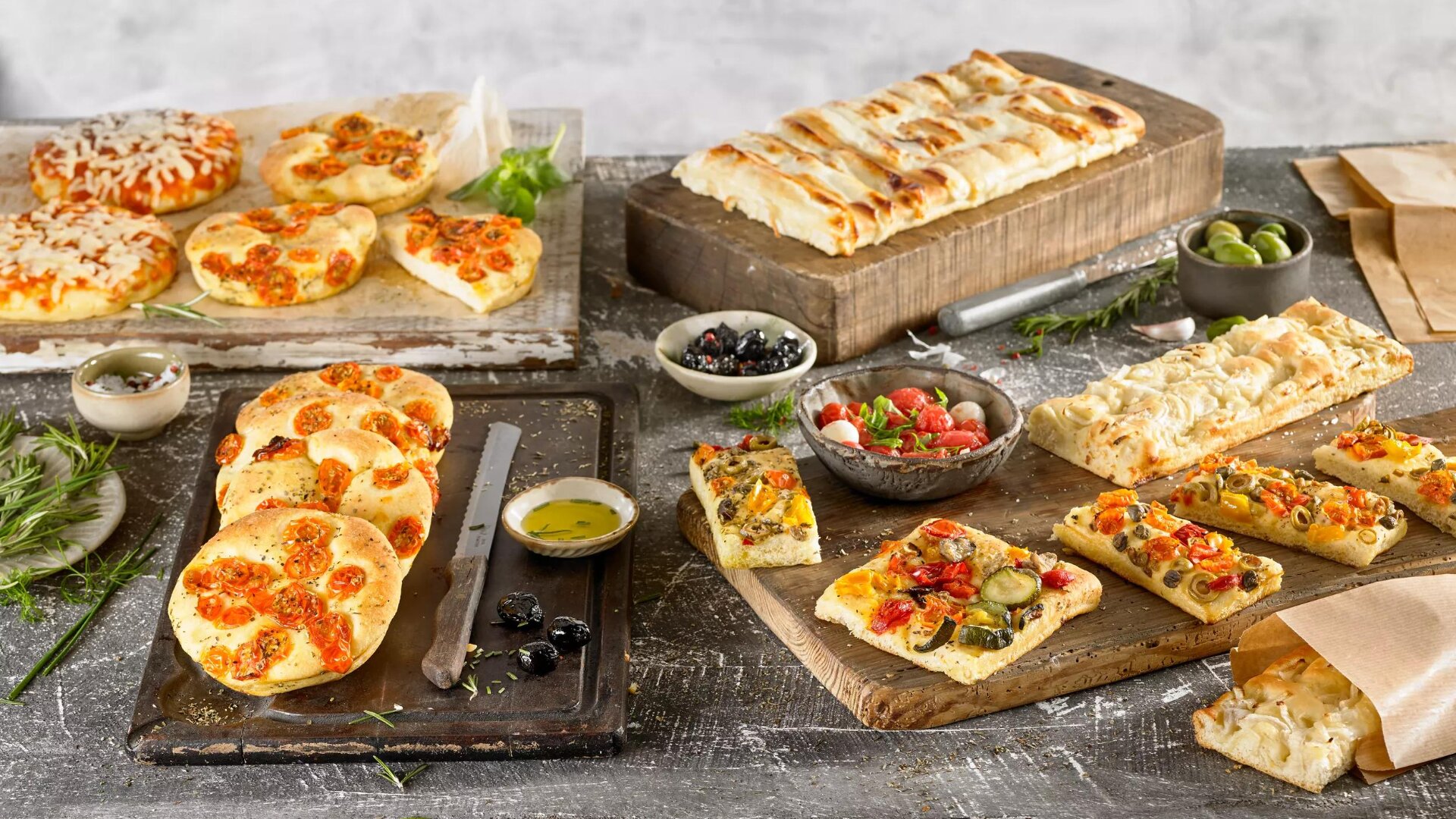 Auf einem rustikalen Tisch stehen verschiedene italienische Brotsorten, darunter Focaccia mit Kirschtomaten, mit Gemüse belegte Fladenbrote, Käsebrötchen, ein geflochtener Laib sowie Schalen mit Oliven und Salat, umgeben von Olivenöl und Kräutern.