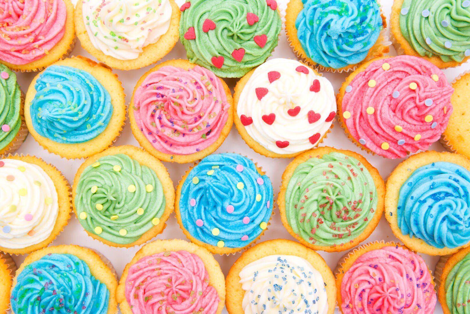 Eine Auswahl an Cupcakes mit buntem Zuckerguss in Rosa, Grün, Blau und Weiß, gekrönt mit verschiedenen Streuseln und herzförmigen Verzierungen, die in einem ordentlichen, überlappenden Muster angeordnet sind.
