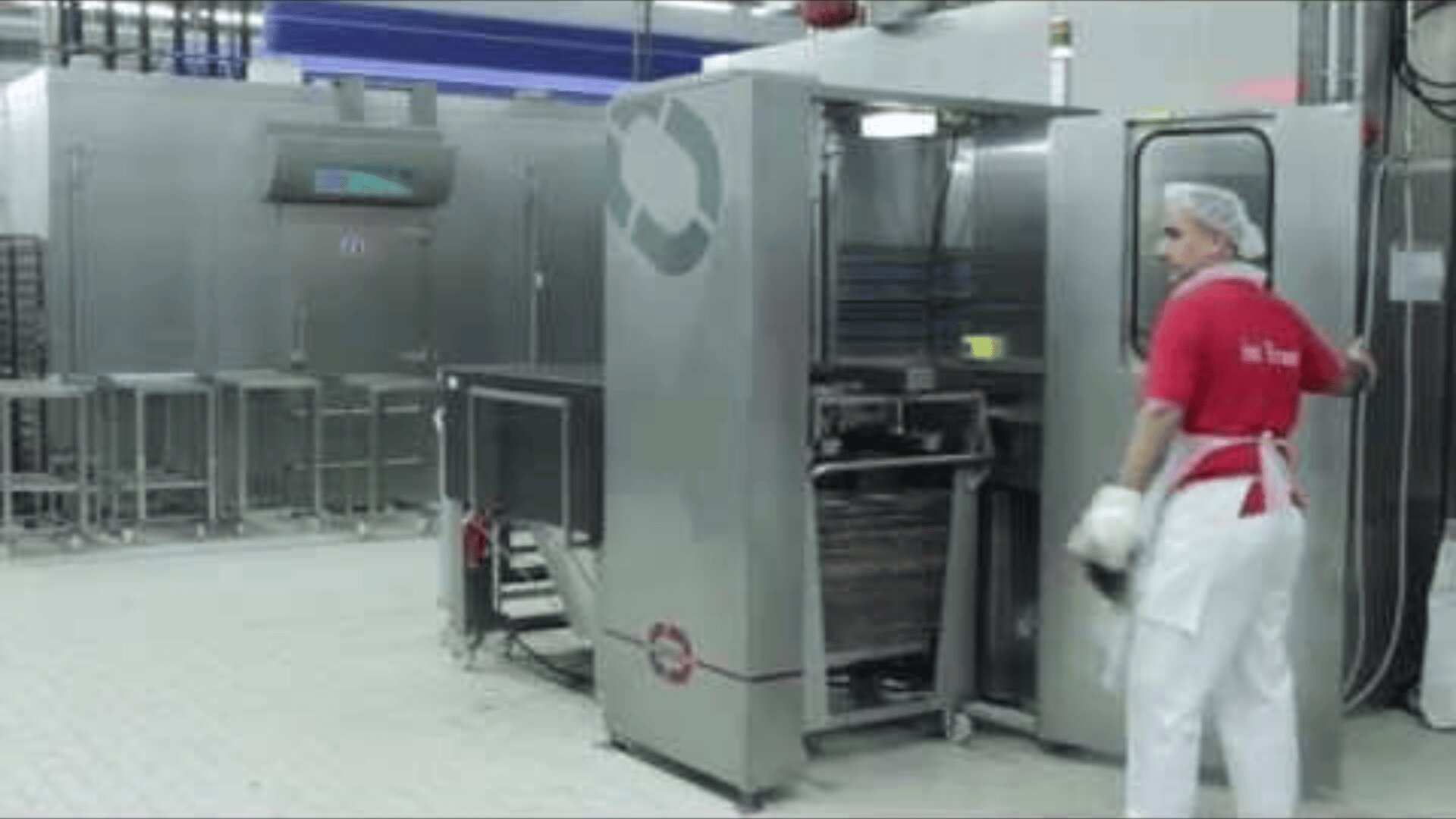 A worker in a red shirt, white apron, and hairnet operates industrial kitchen equipment in a clean, modern food processing facility. Stainless steel machines and tiled floors are visible.