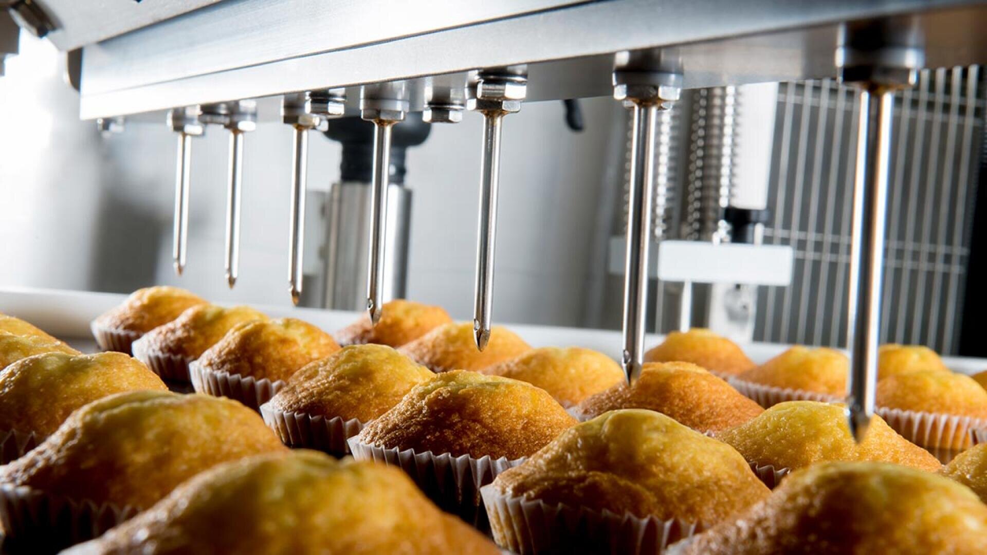 A close-up of a food production machine injecting filling into rows of golden-brown muffins, each placed in paper liners, inside a factory setting.