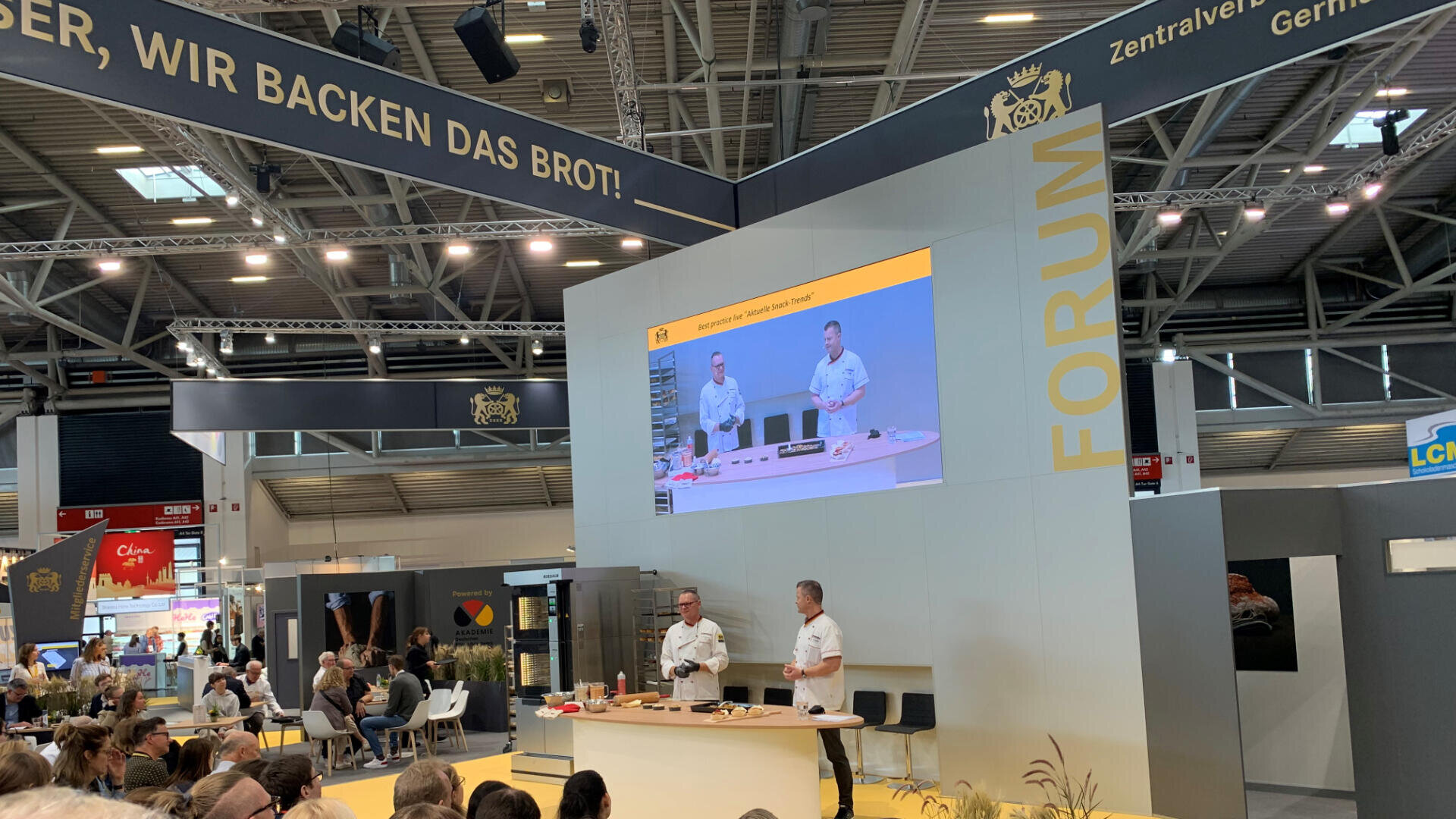 Zwei Köche geben auf einer Messebühne eine Backvorführung. Ein großer Bildschirm zeigt dem sitzenden Publikum ihre Aktionen. Darüber hängt ein Schild mit der Aufschrift „Wir backen das Brot!“. Gelb-graues Dekor mit Forum-Logo.