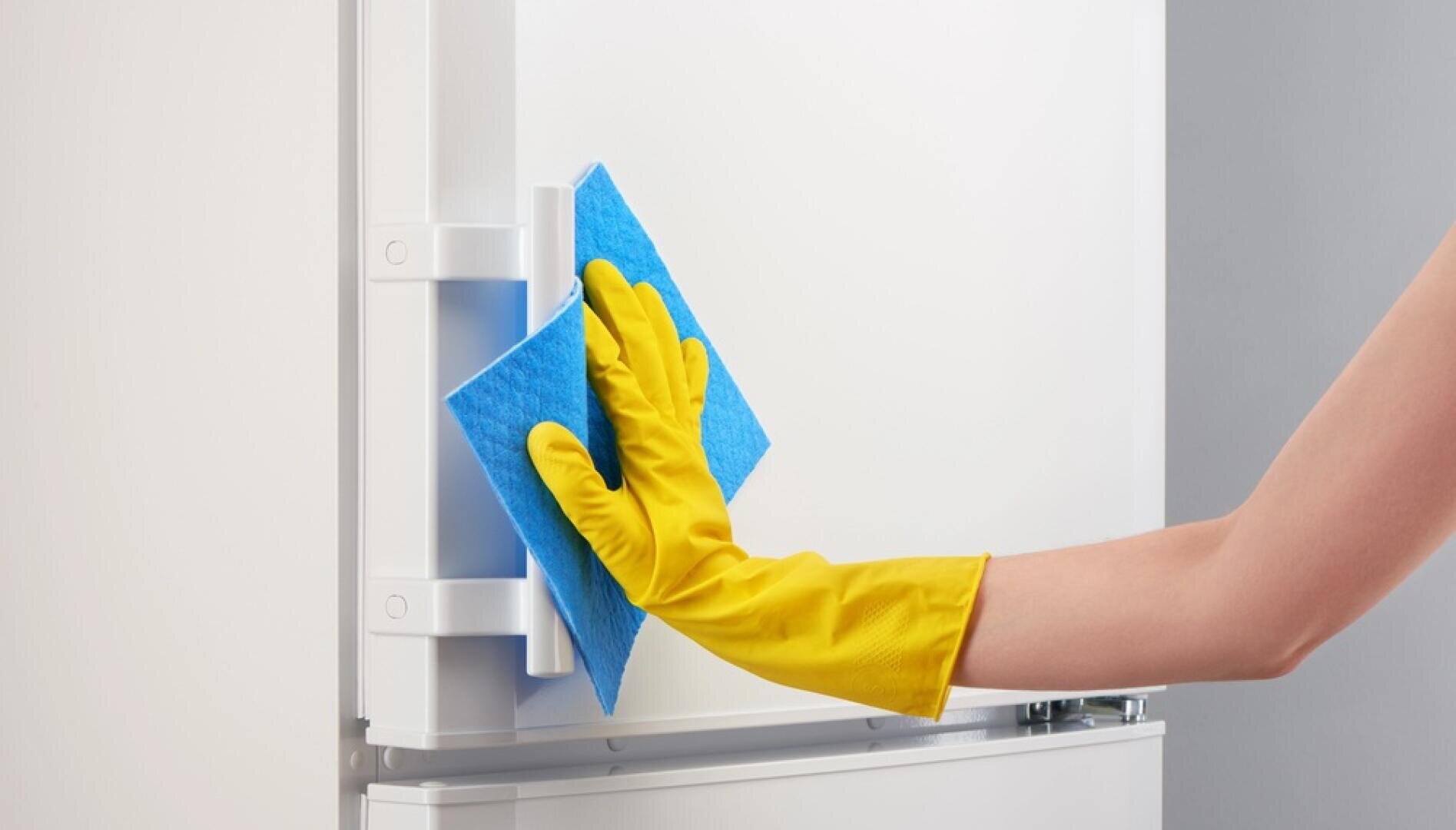A person wearing a yellow rubber glove cleans a white refrigerator door with a blue sponge.