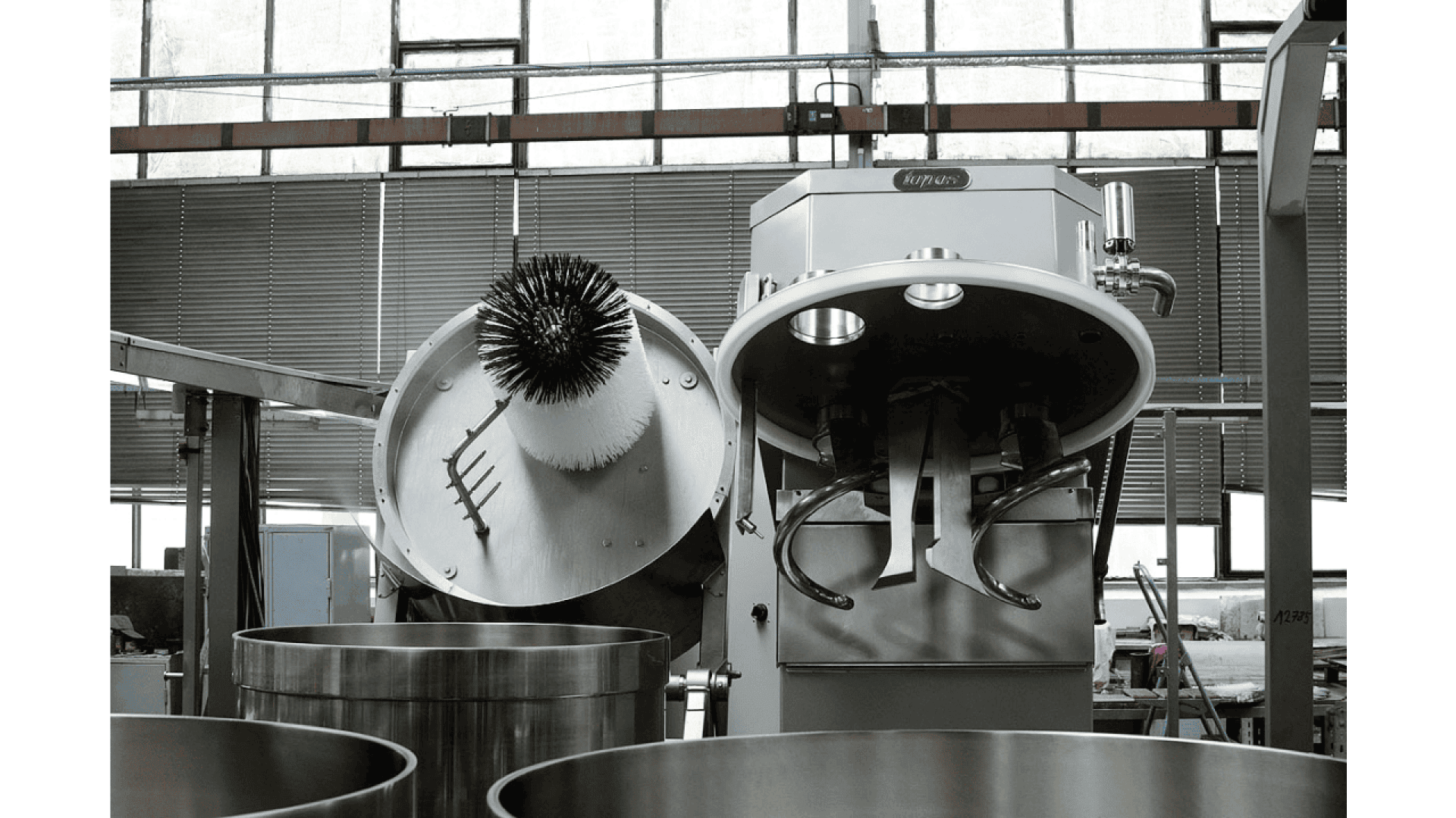 Industrielle Küchenmaschine mit geöffnetem Deckel, der die Mischarme und eine Bürste im Inneren des Deckels zeigt. Vorne stehen große Edelstahl-Mischschüsseln, und im Hintergrund sind Fenster und Industrieanlagen zu sehen.