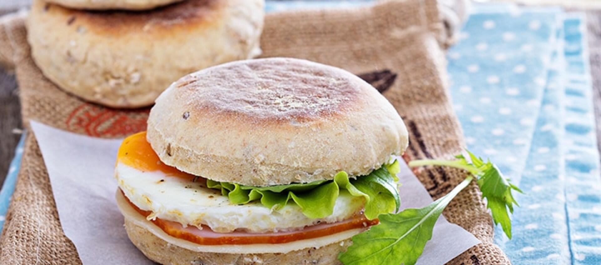 A breakfast sandwich on a whole wheat English muffin with egg, cheese, lettuce, and tomato, placed on parchment paper with a sprig of parsley nearby. Another sandwich is slightly blurred in the background.