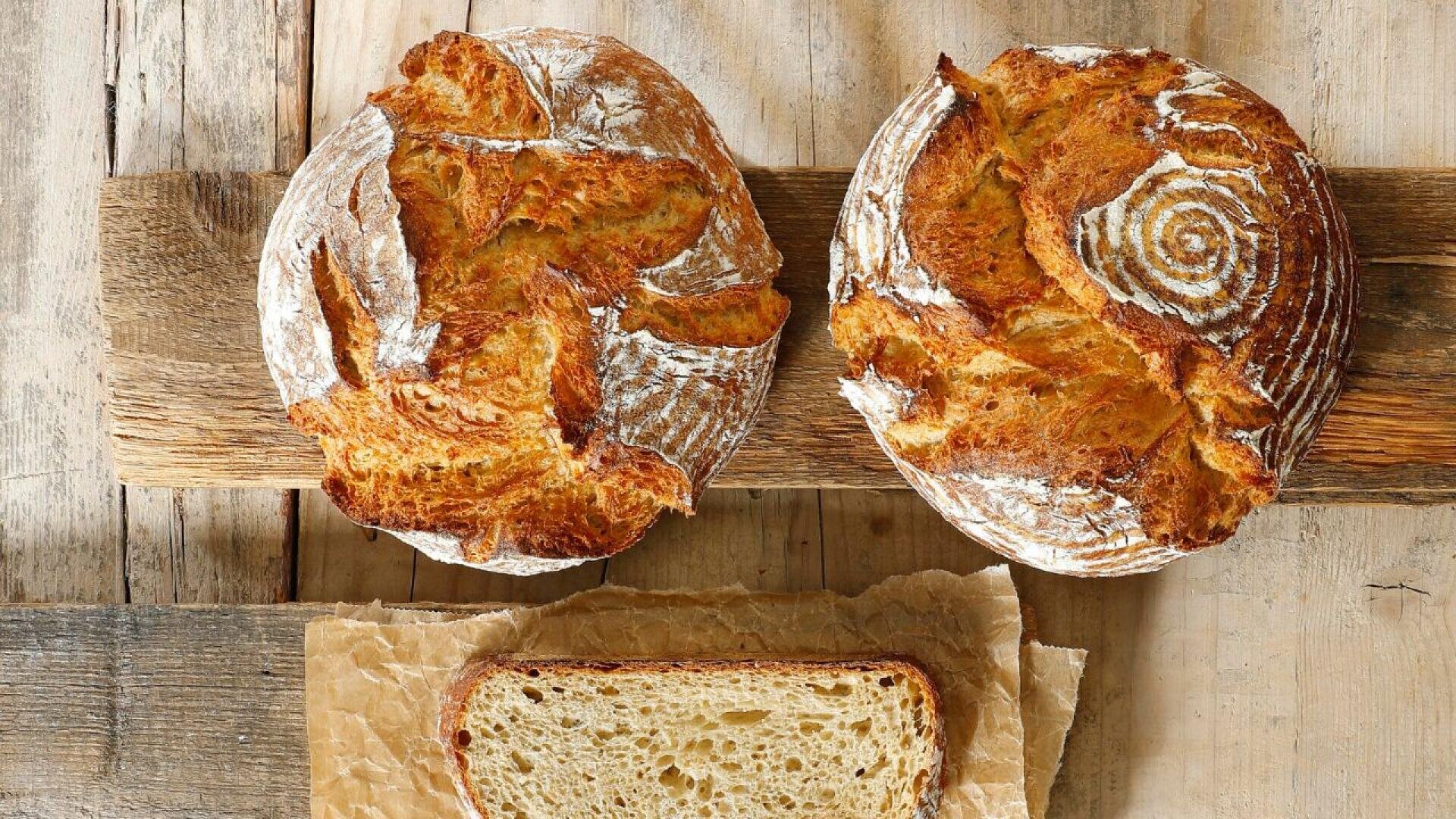 Zwei runde Laibe rustikales Handwerkerbrot mit goldener Kruste und dekorativem Mehlmuster liegen auf einem Holzbrett über einer dicken Brotscheibe auf braunem Pergamentpapier.