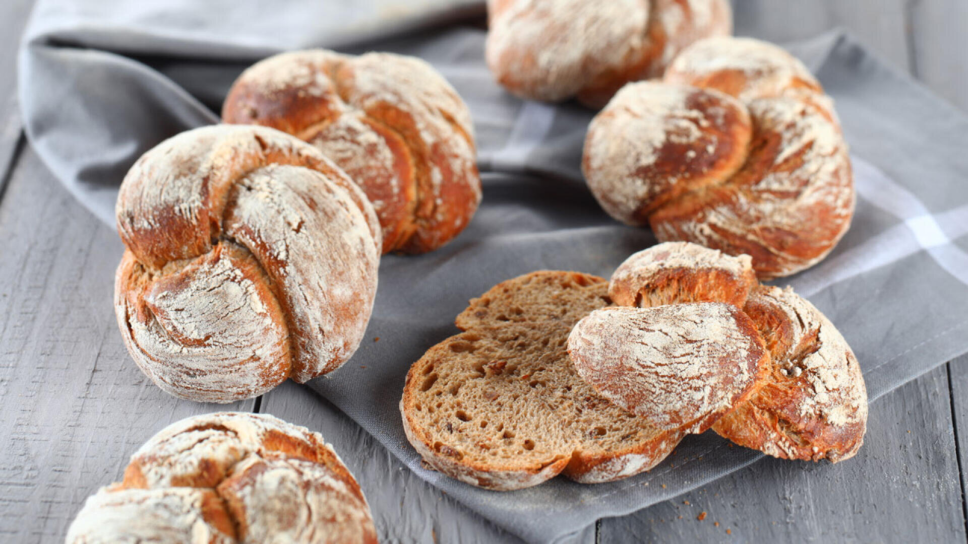 Mehrere runde, rustikale, mit Mehl bestäubte Brötchen liegen auf einem grauen Tuch über einer Holzfläche. Ein Brötchen ist aufgeschnitten und zeigt sein weiches, braunes Inneres.