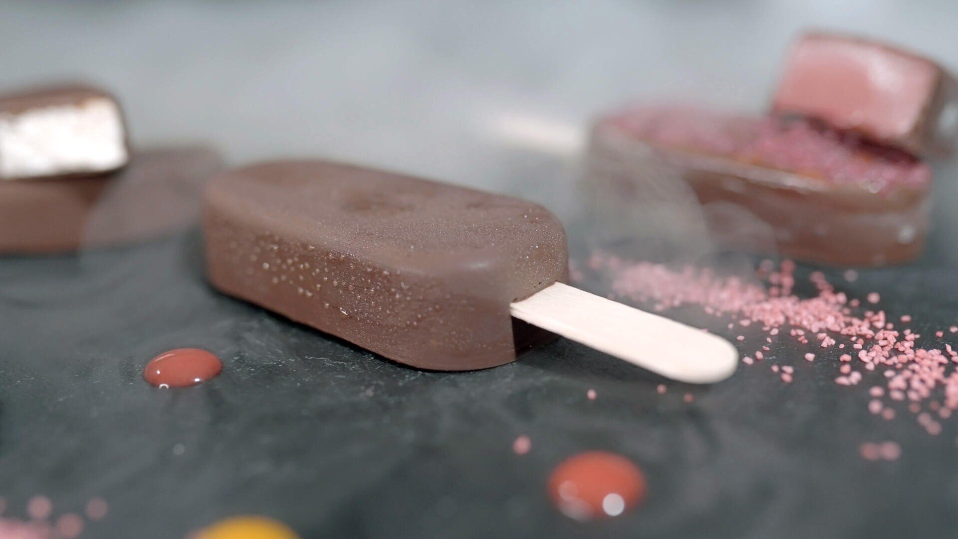 Ein schokoladenüberzogener Eisriegel am Stiel ruht auf einer dunklen Fläche, umgeben von roten und gelben Siruptropfen und rosa Streuseln, mit einem verschwommenen Dessert im Hintergrund.