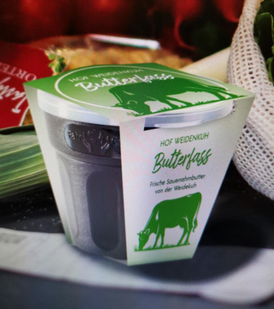 Ein Behälter mit frischer Sauerrahmbutter von Butterfass vom Hof Weidenkuh mit einer grün-weißen Verpackung, auf der eine Kuh abgebildet ist, steht auf einem Tisch neben frischem Gemüse.
