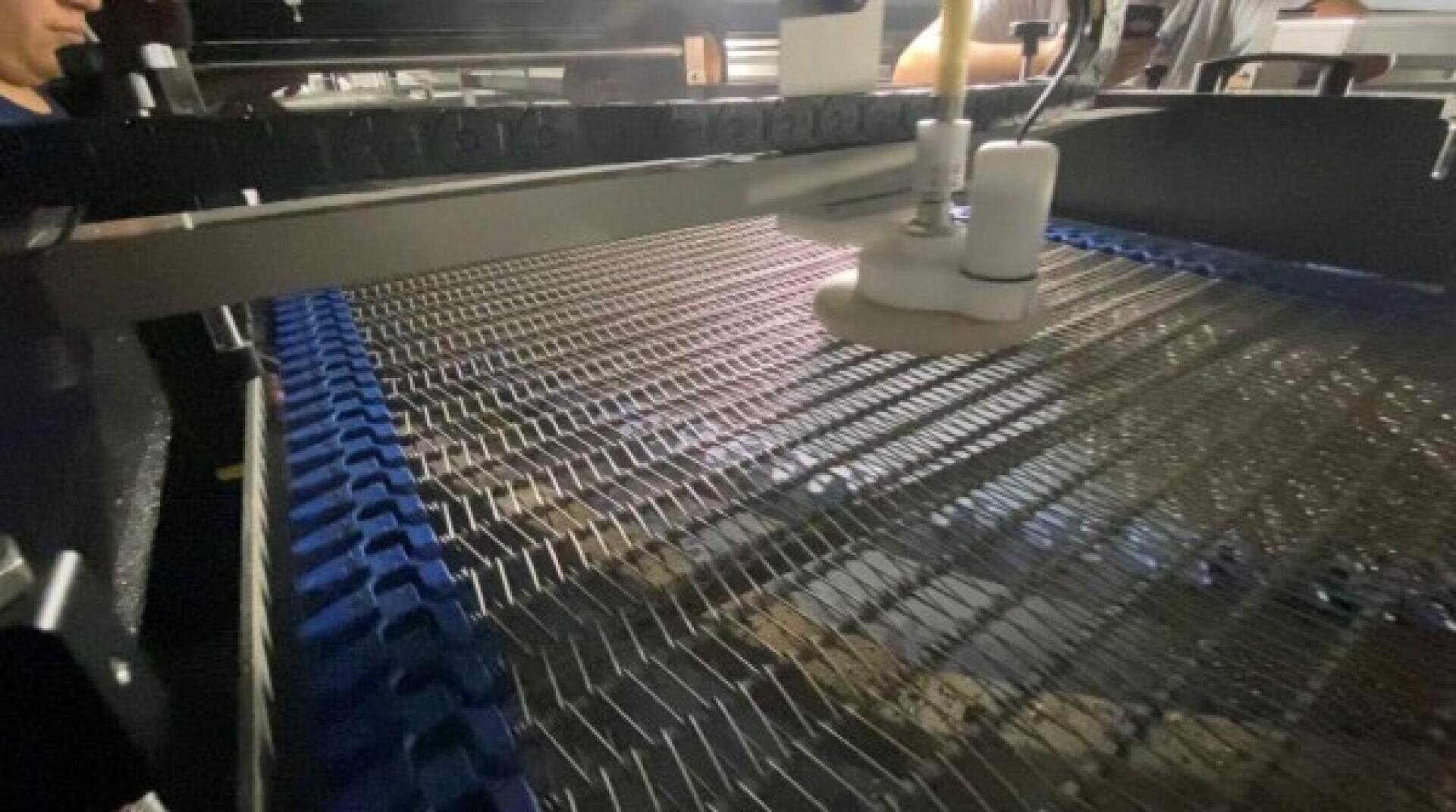 A close-up view of an industrial conveyor belt system with metal mesh and blue edges, featuring a white robotic arm or tool suspended above the belt.