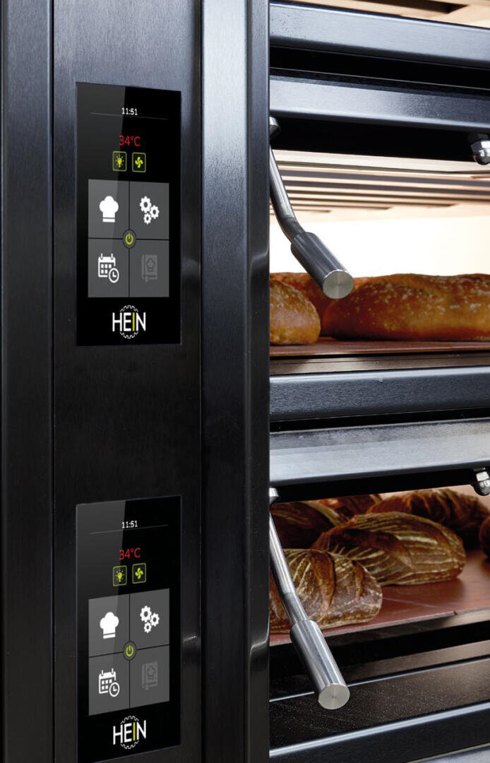 Nahaufnahme der elektronischen Schalttafeln eines kommerziellen Backofens, wobei die Brotlaibe in den offenen Ofenkammern sichtbar backen. Die Kontrollbildschirme zeigen Temperatur- und Backsymbole an.