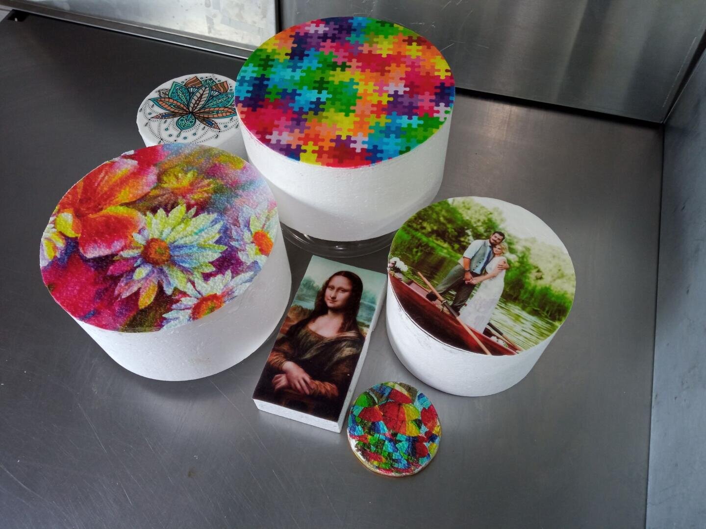 Five decorated cakes with printed edible images, including colorful patterns, flowers, a puzzle design, a bride and groom in a boat, a mandala, and the Mona Lisa, arranged on a stainless steel surface.