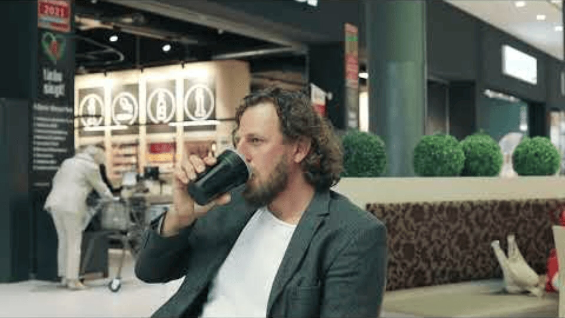 A bearded man with curly hair, wearing a gray blazer and white shirt, sits in a mall or shopping center, sipping from a black cup. In the background, a person pushes a shopping cart near store displays.