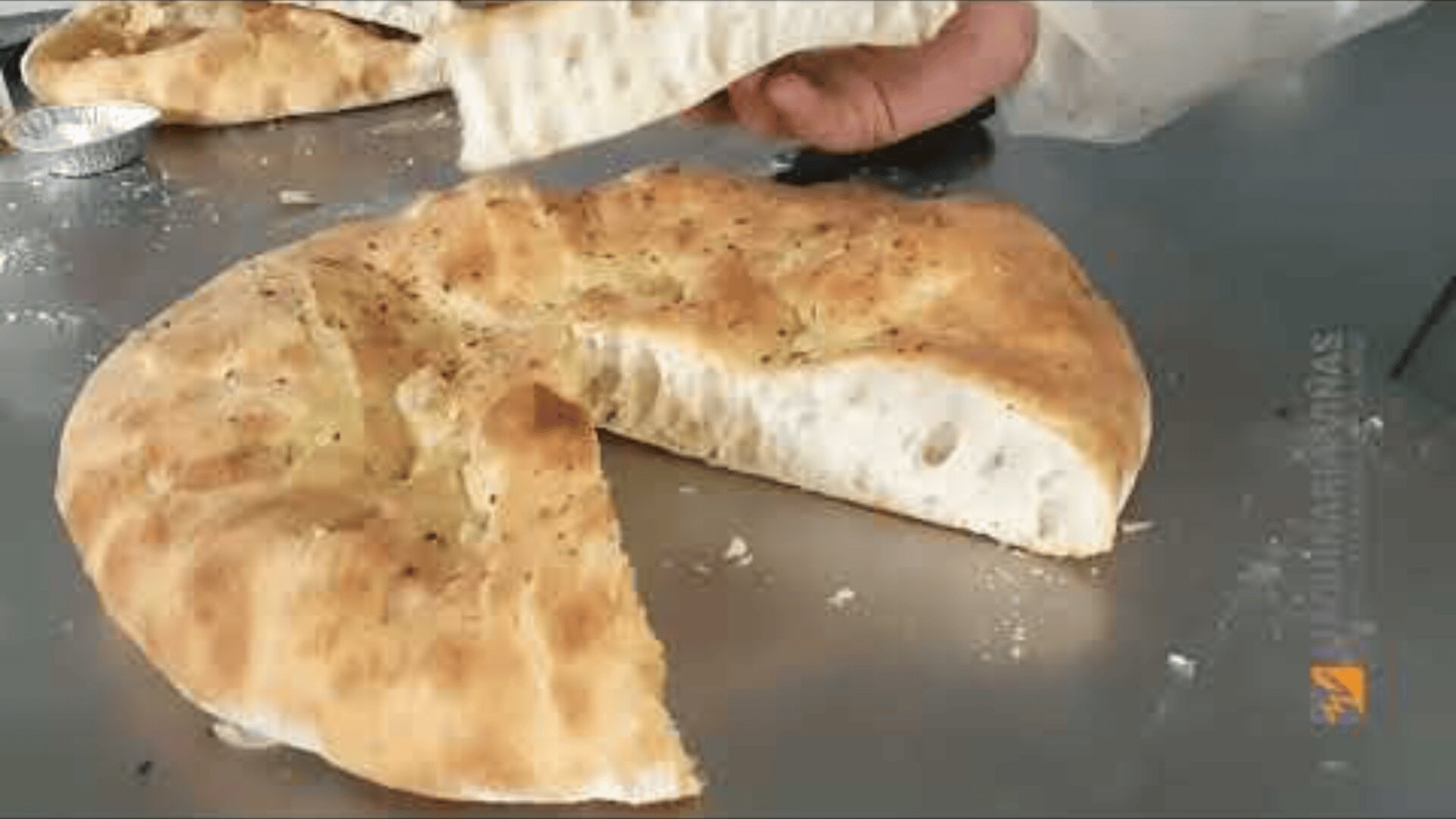 One hand holds a slice of round, golden-brown focaccia bread, which has been cut open to reveal its airy, soft interior. The bread lies on a metal surface, lightly sprinkled with crumbs.