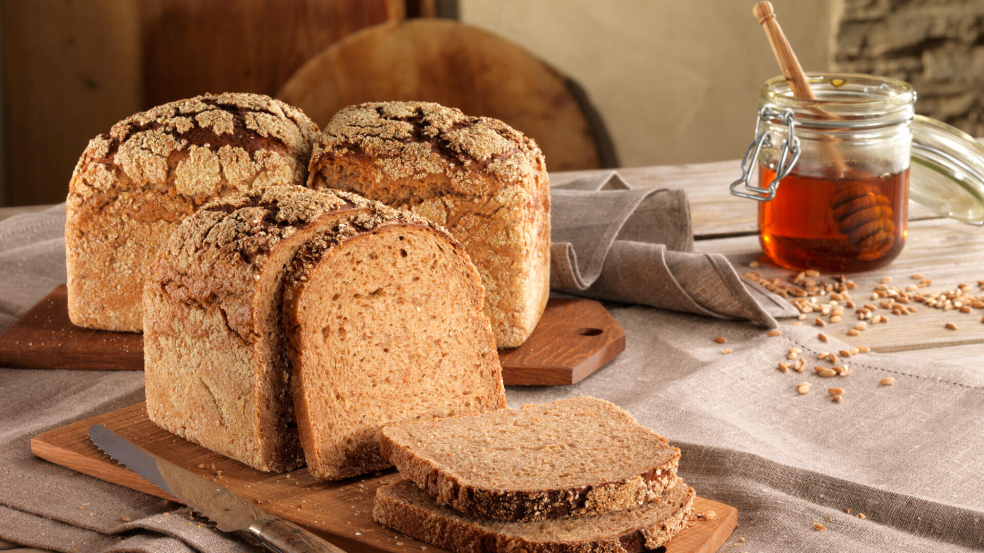 Drei Laibe rustikales Vollkornbrot und mehrere Scheiben liegen auf Holzbrettern neben einem Messer, im Hintergrund auf einem mit Leinen gedeckten Tisch ein Glas Honig und ein Honiglöffel.