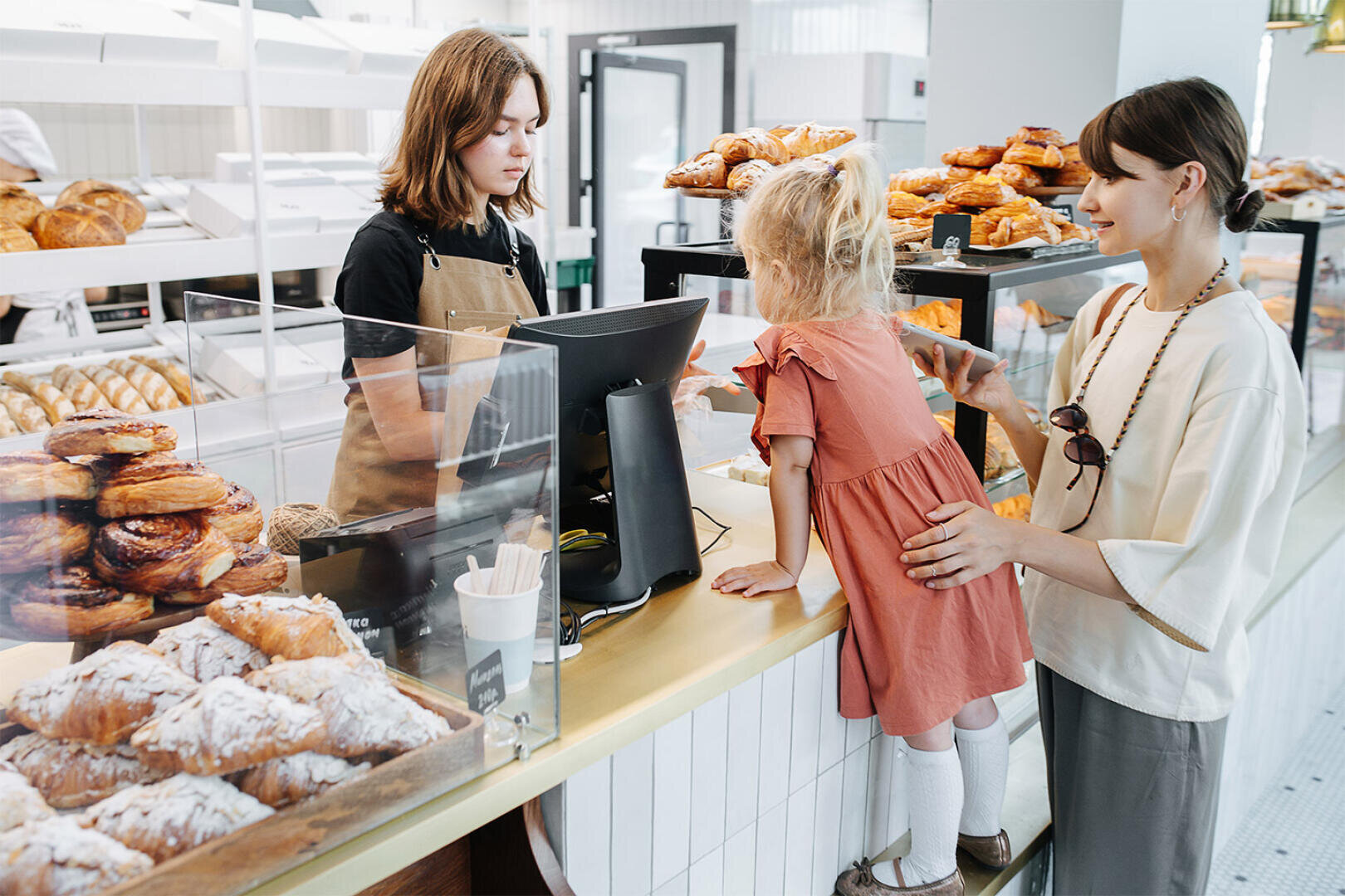 Eine junge Frau und ein Kind stehen an der Theke einer Bäckerei und suchen sich Gebäck aus. Eine Mitarbeiterin in Schürze hilft ihnen dabei. Die Vitrinen sind mit verschiedenen Backwaren gefüllt.