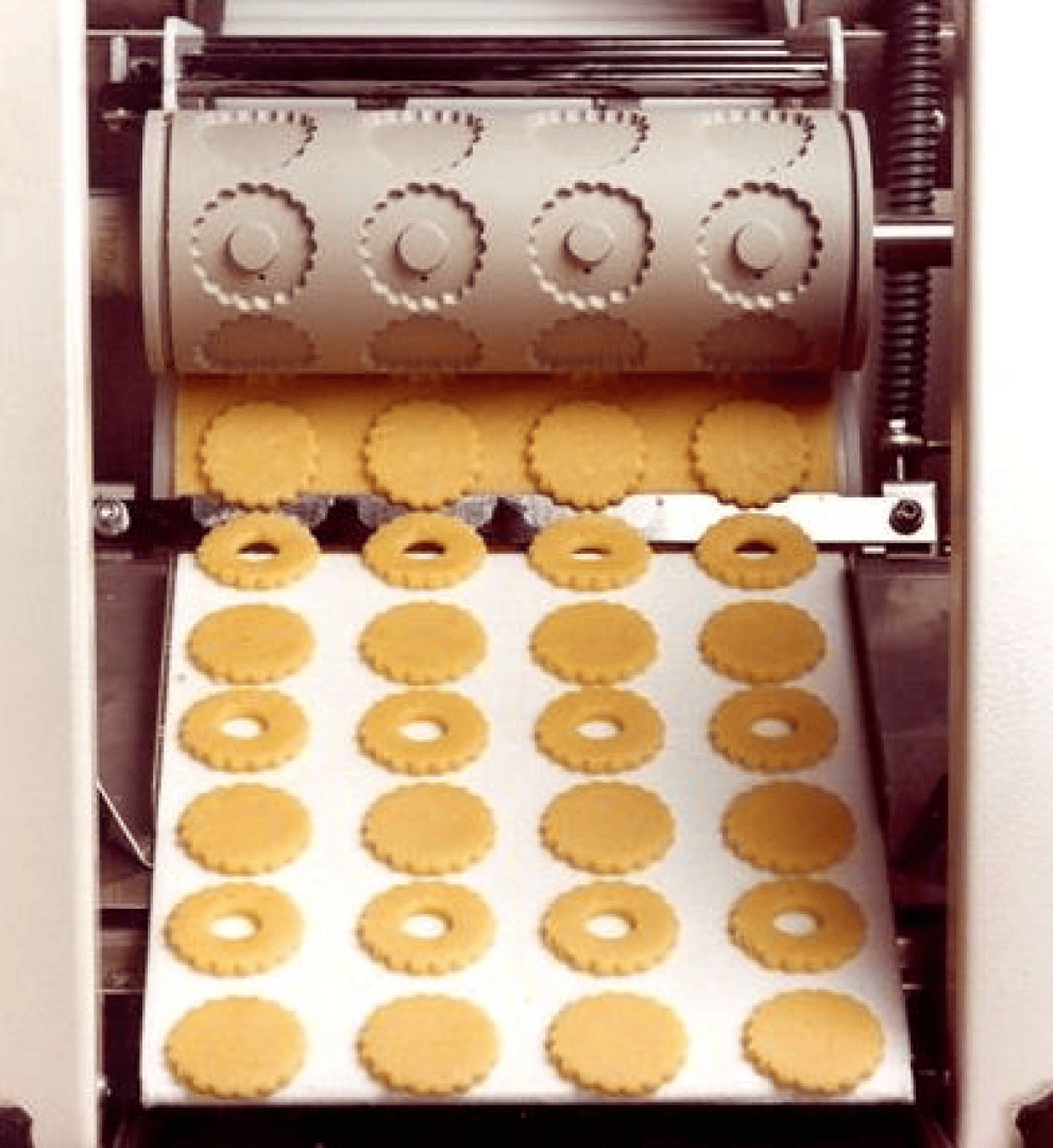 Eine Maschine stanzt runde, gezackte Kekse mit Löchern in der Mitte auf ein Förderband und produziert gleichmäßige Reihen von Rohteigformen.