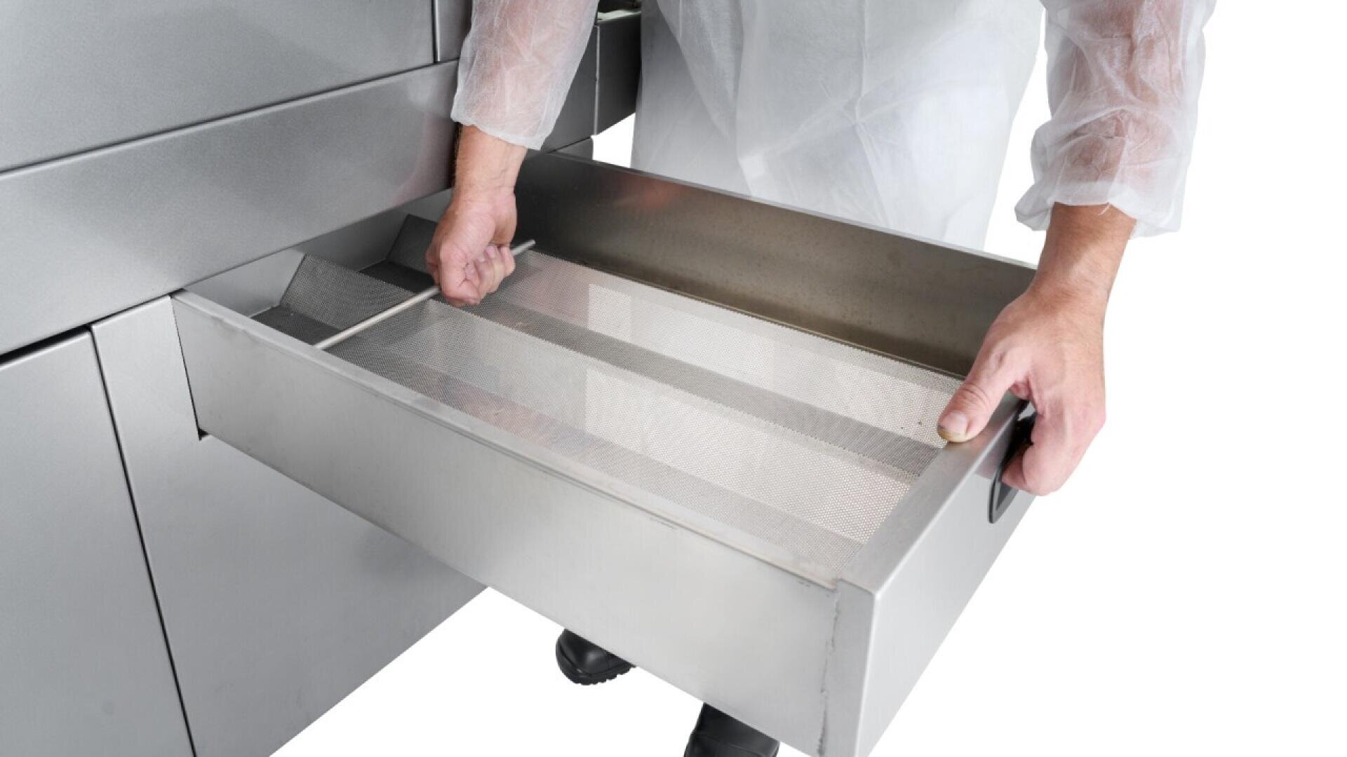 A person wearing protective sleeves pulls out a stainless steel mesh drawer from an industrial machine, likely used for food processing or filtration.