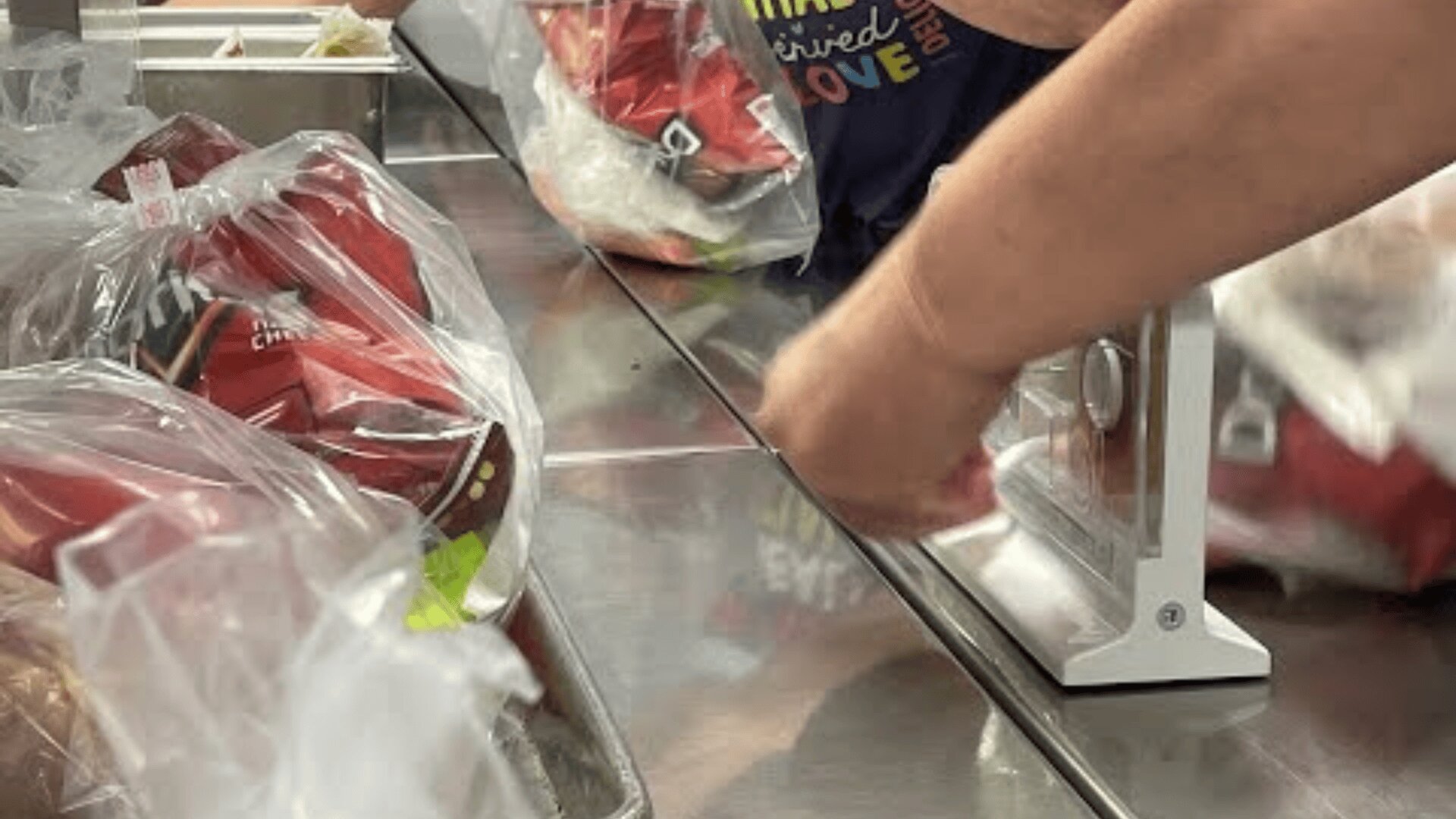 Close-up of hands sealing bags of packaged food with a heat sealer on a metal counter. Several plastic bags filled with small red snack packs are visible.