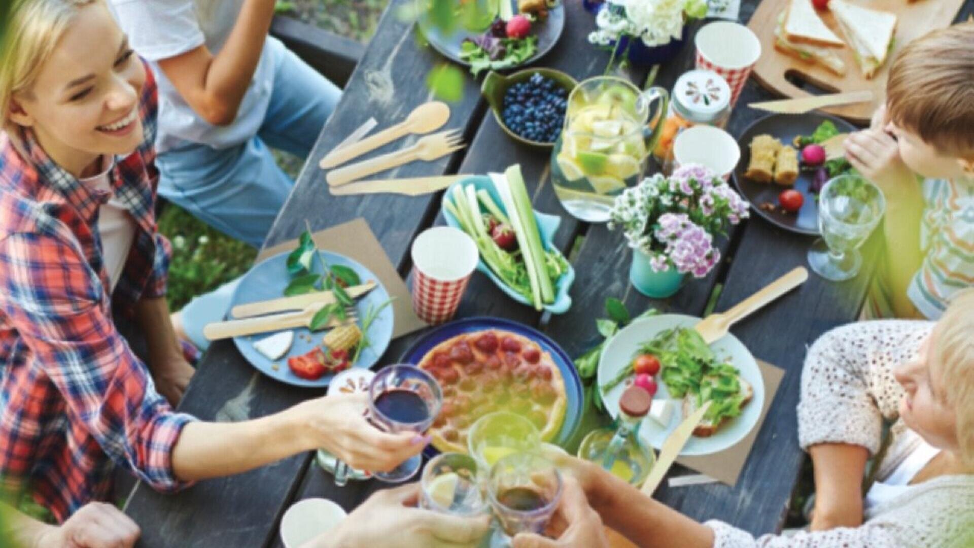 Eine Gruppe von Menschen sitzt an einem Tisch im Freien und teilt eine Mahlzeit mit Salaten, Obst und Getränken und stößt mit Gläsern an. Der Tisch ist mit Blumen und buntem Geschirr dekoriert, was eine fröhliche, festliche Atmosphäre schafft.
