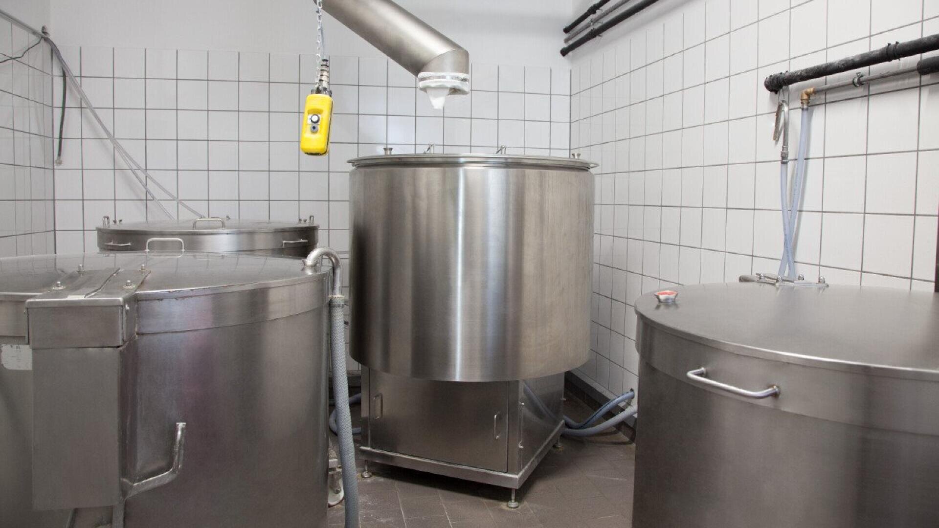 Stainless steel industrial tanks with lids and pipes in a clean, white-tiled room, likely used for food or beverage processing. A yellow control device hangs from the ceiling.