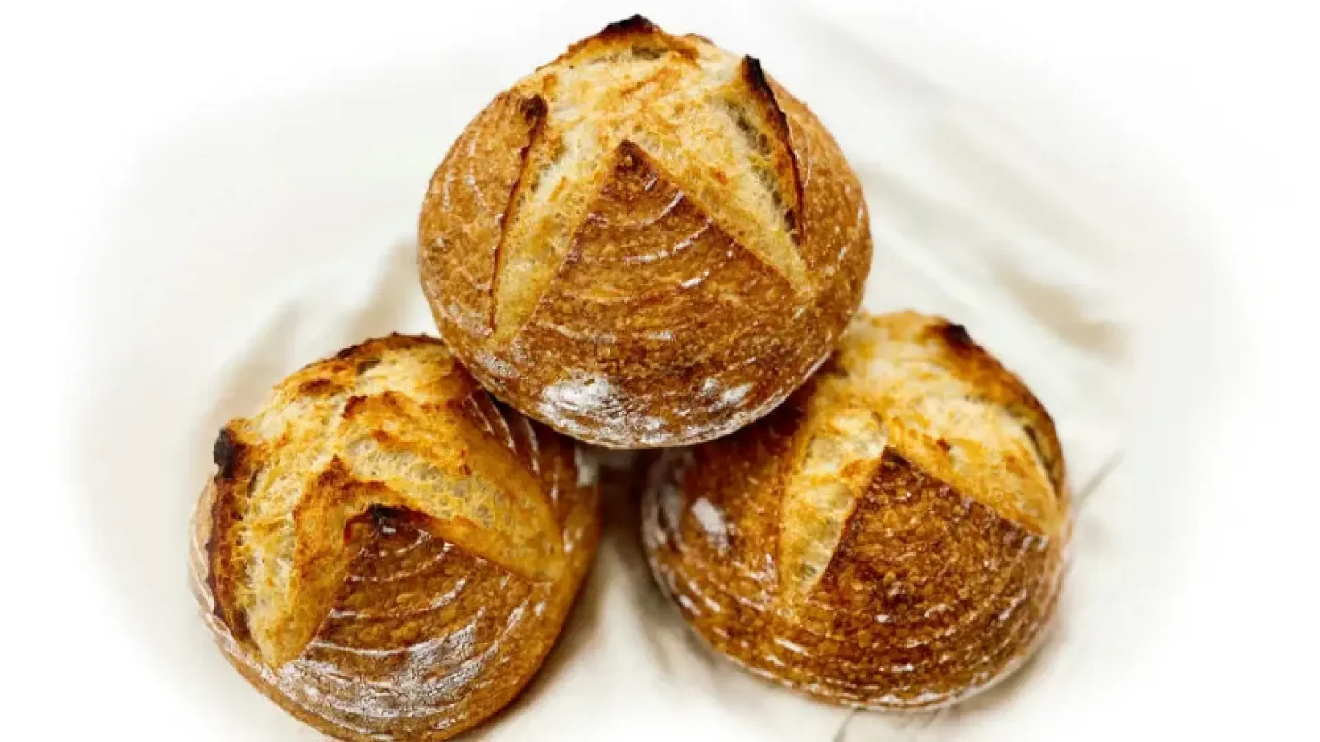 Three round, golden-brown sourdough bread loaves with rustic, crusty tops sit on a white cloth background. Each loaf has a distinct scoring pattern and a lightly floured surface.
