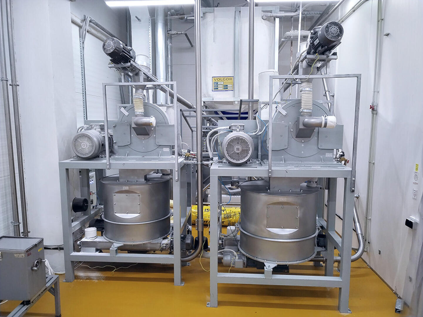 Zwei große Industriemaschinen mit Motoren, Ventilatoren und zylindrischen Behältern in einer sauberen, hell erleuchteten Halle mit weißen Wänden und gelbem Boden. Verschiedene Rohre und Kabel verbinden die Geräte.