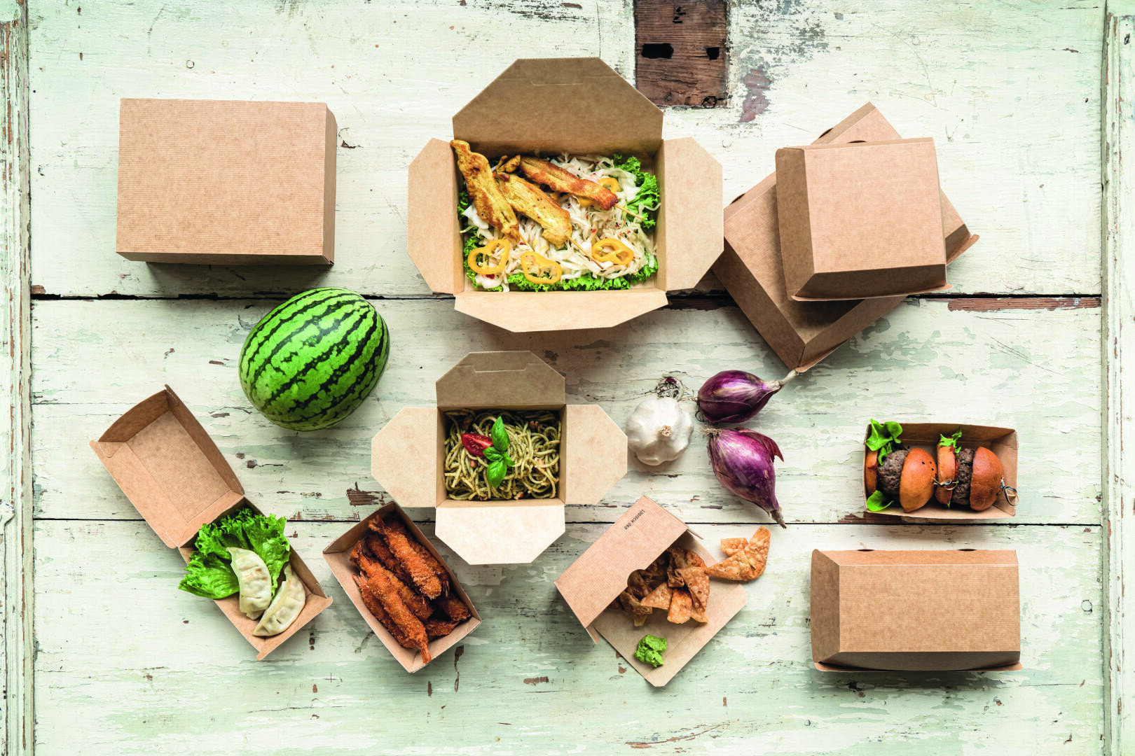 Assorted takeout food in open and closed brown paper boxes, with a small whole watermelon, purple onions, and garlic, all arranged on a rustic white wooden surface.