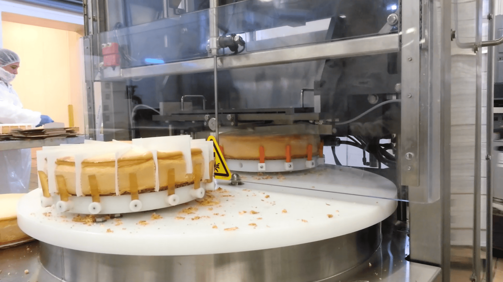 A cheesecake is being sliced by an automated machine in a commercial kitchen. A worker wearing a hairnet and mask is visible in the background. Crumbs are scattered on the round cutting surface.