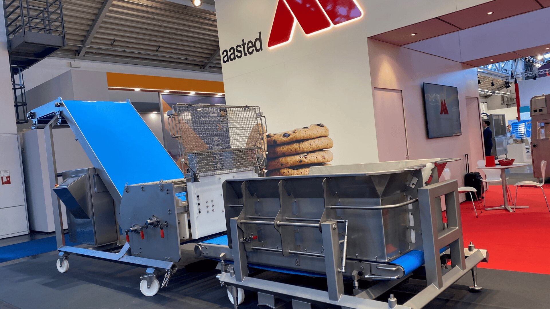 A large industrial food processing machine with a conveyor belt and a stack of oversized cookies on display at an exhibition booth with red and white decor and the aasted logo on the wall.