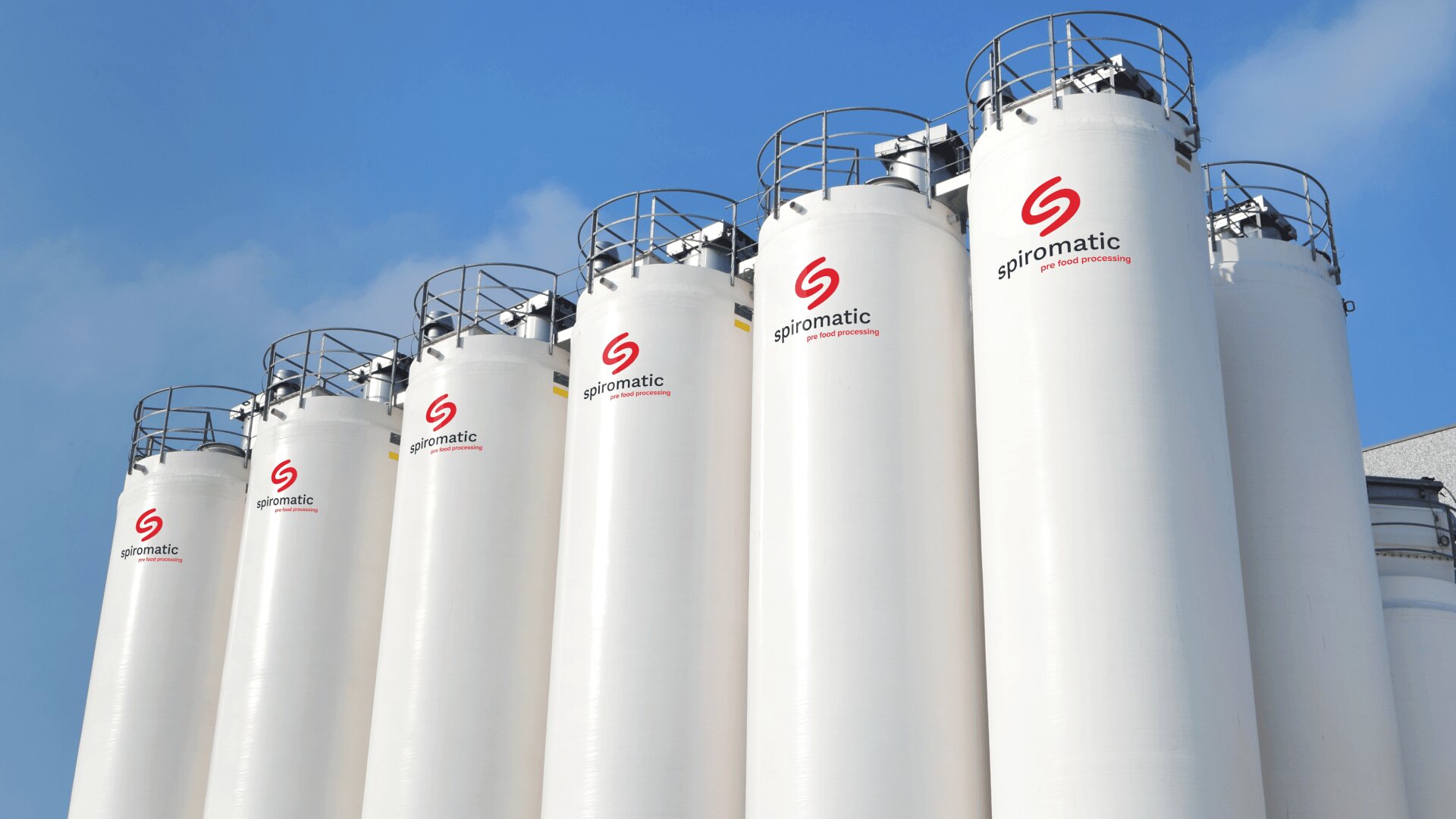 A row of large white industrial silos with red Spiromatic logos and safety railings on top, set against a blue sky.